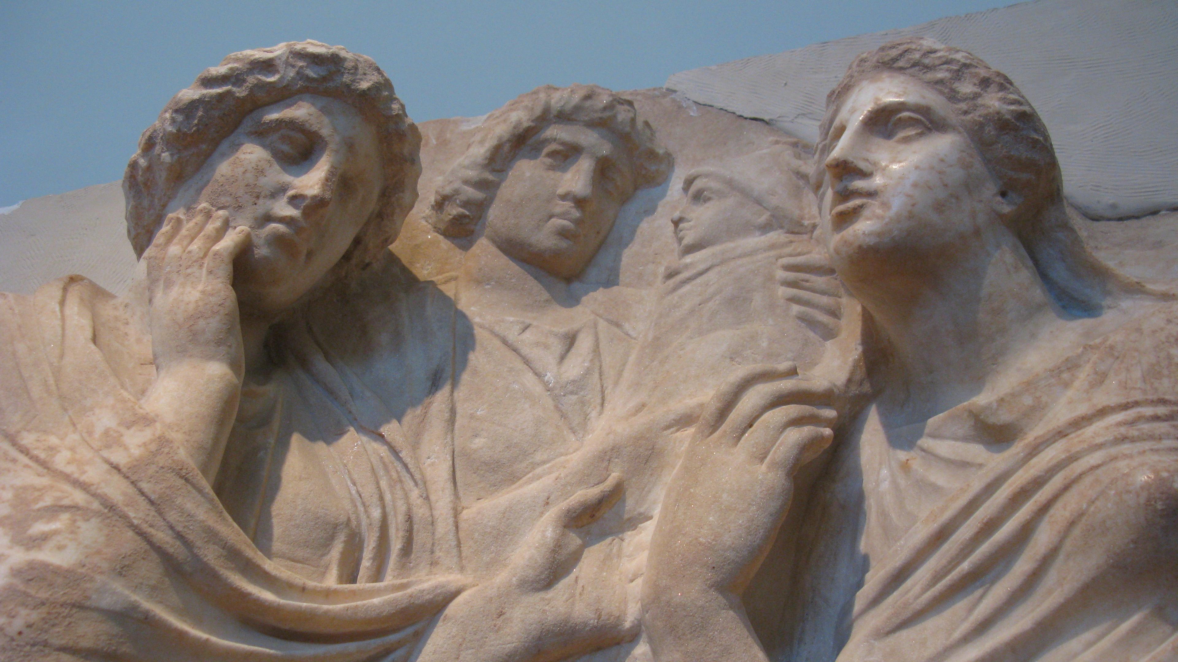 Photo of an ancient stone relief depicting four figures in draped garments with expressive faces, shown in close-up.