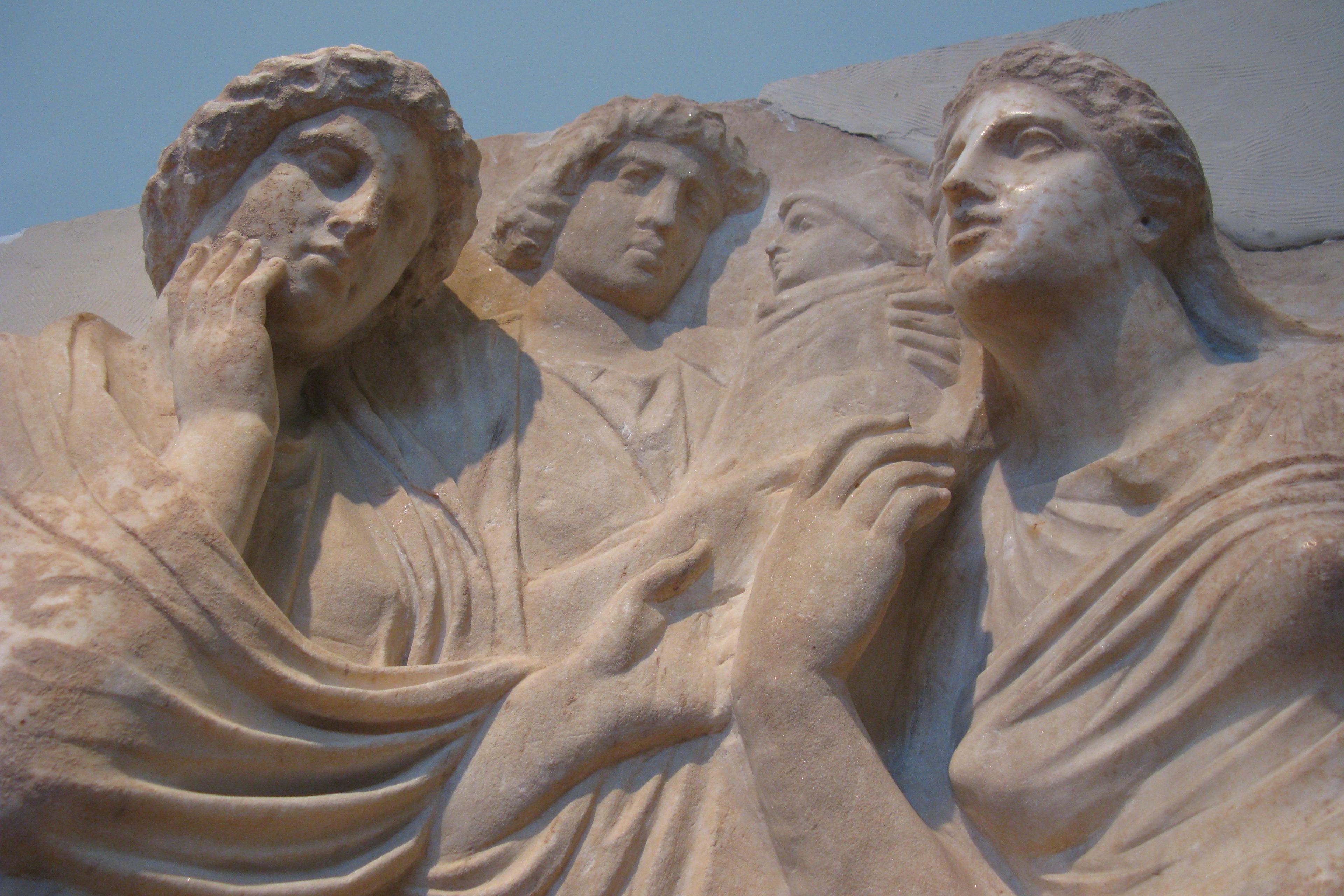 Photo of an ancient stone relief depicting four figures in draped garments with expressive faces, shown in close-up.