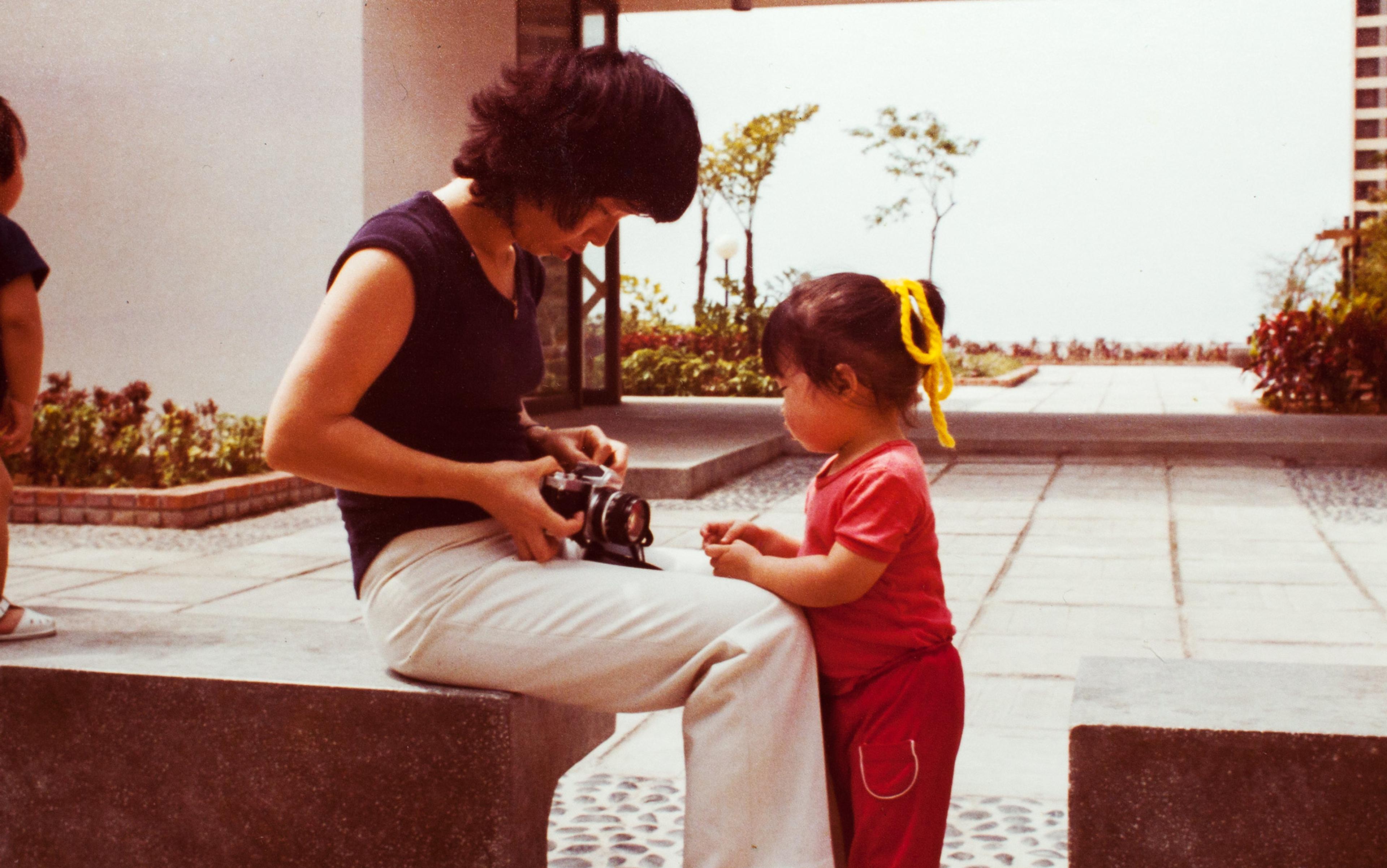 A vintage photo of a woman showing a young girl a camera while sitting on a concrete bench in an outdoor setting.