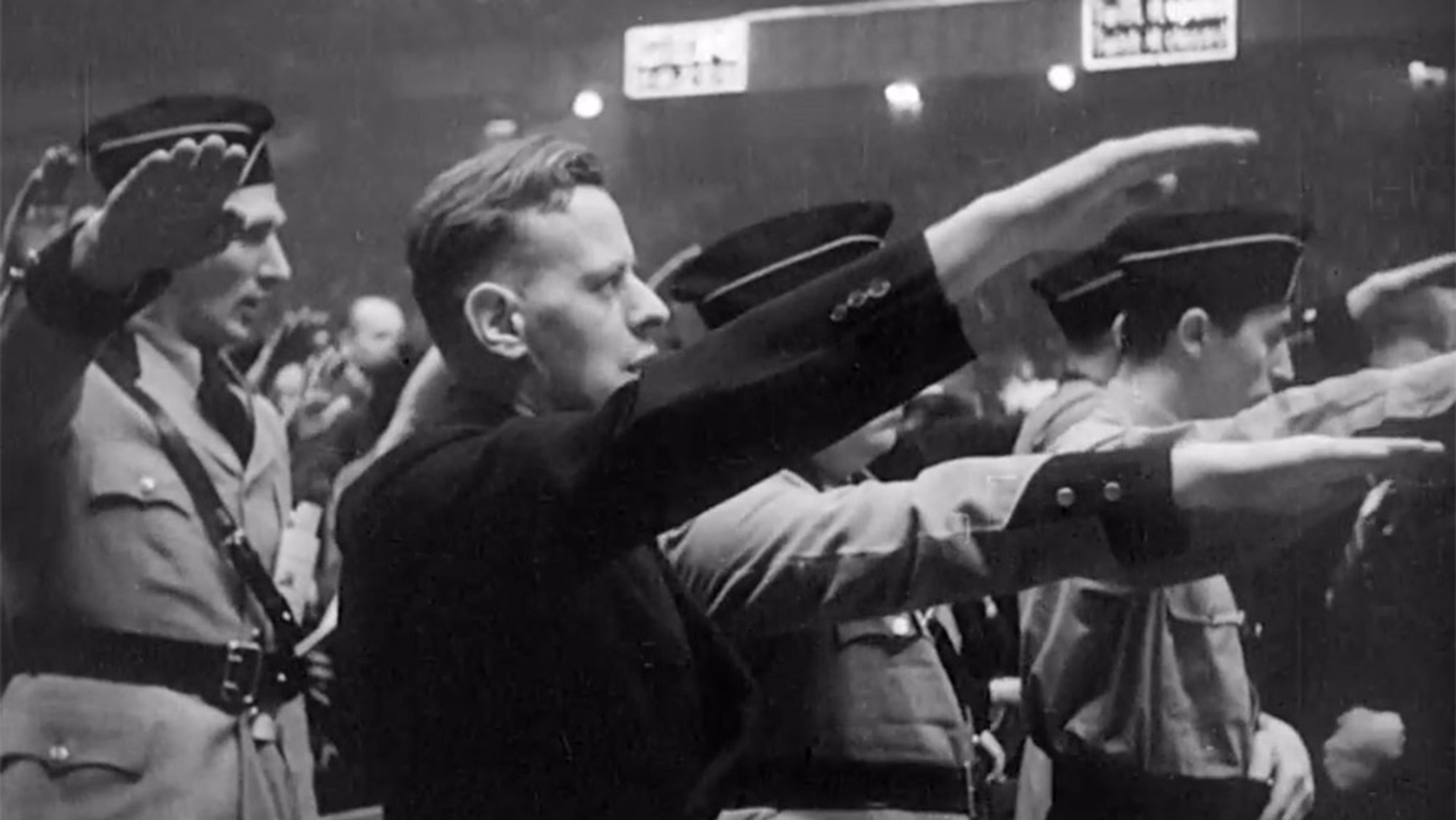 Black and white photo of men in uniform saluting with outstretched arms, standing in a formation at an indoor event or gathering.