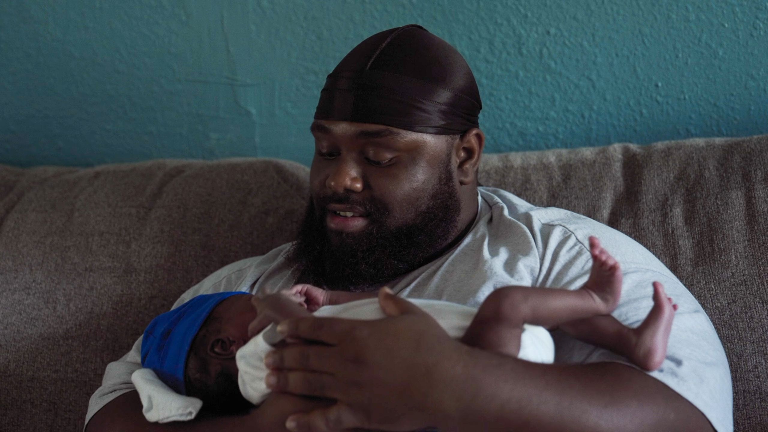 Photo of a man in a black cap holding a baby in white, with a blue cap, sitting on a grey couch against a blue wall.