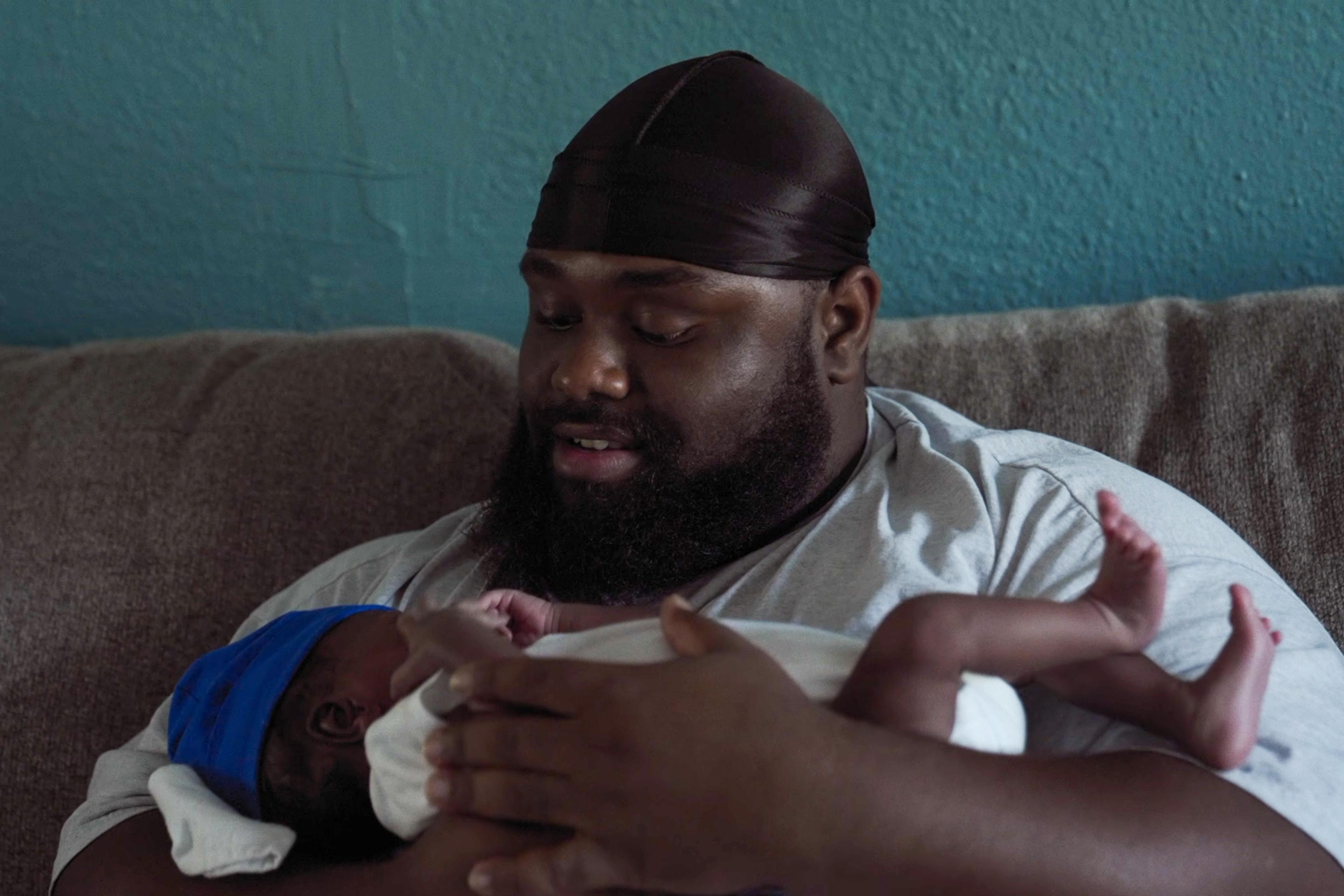 Photo of a man in a black cap holding a baby in white, with a blue cap, sitting on a grey couch against a blue wall.