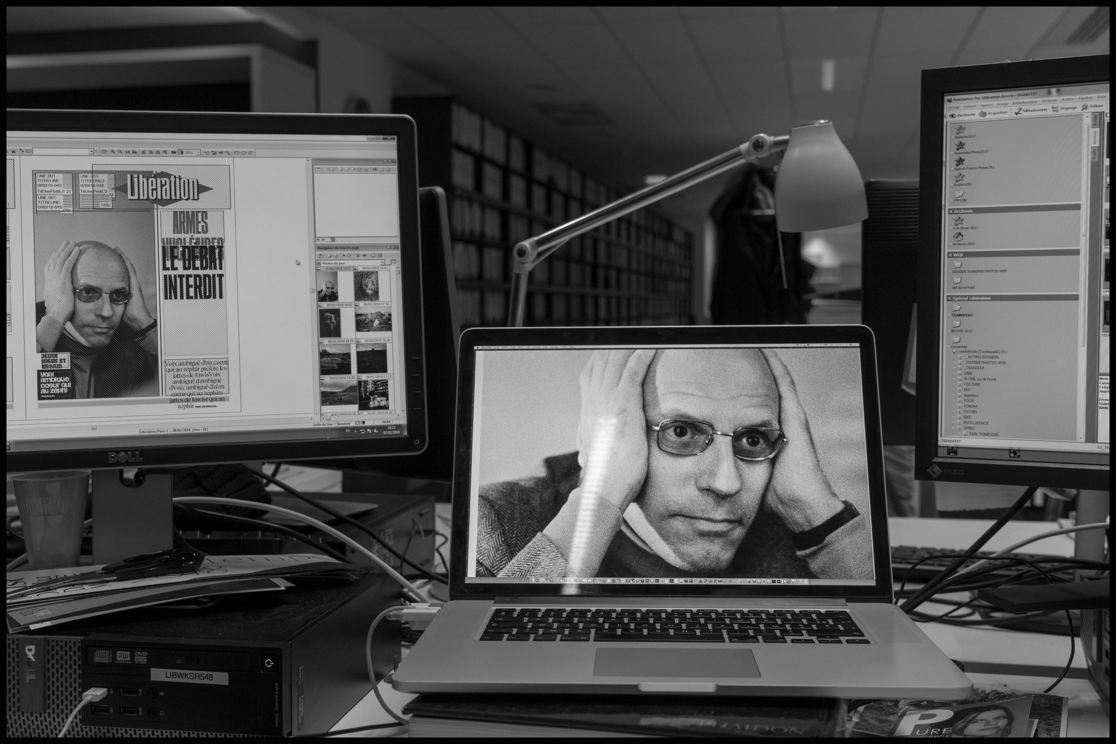 Black and white photo of an office with a laptop and monitors, showing an image of a person with hands on their head.