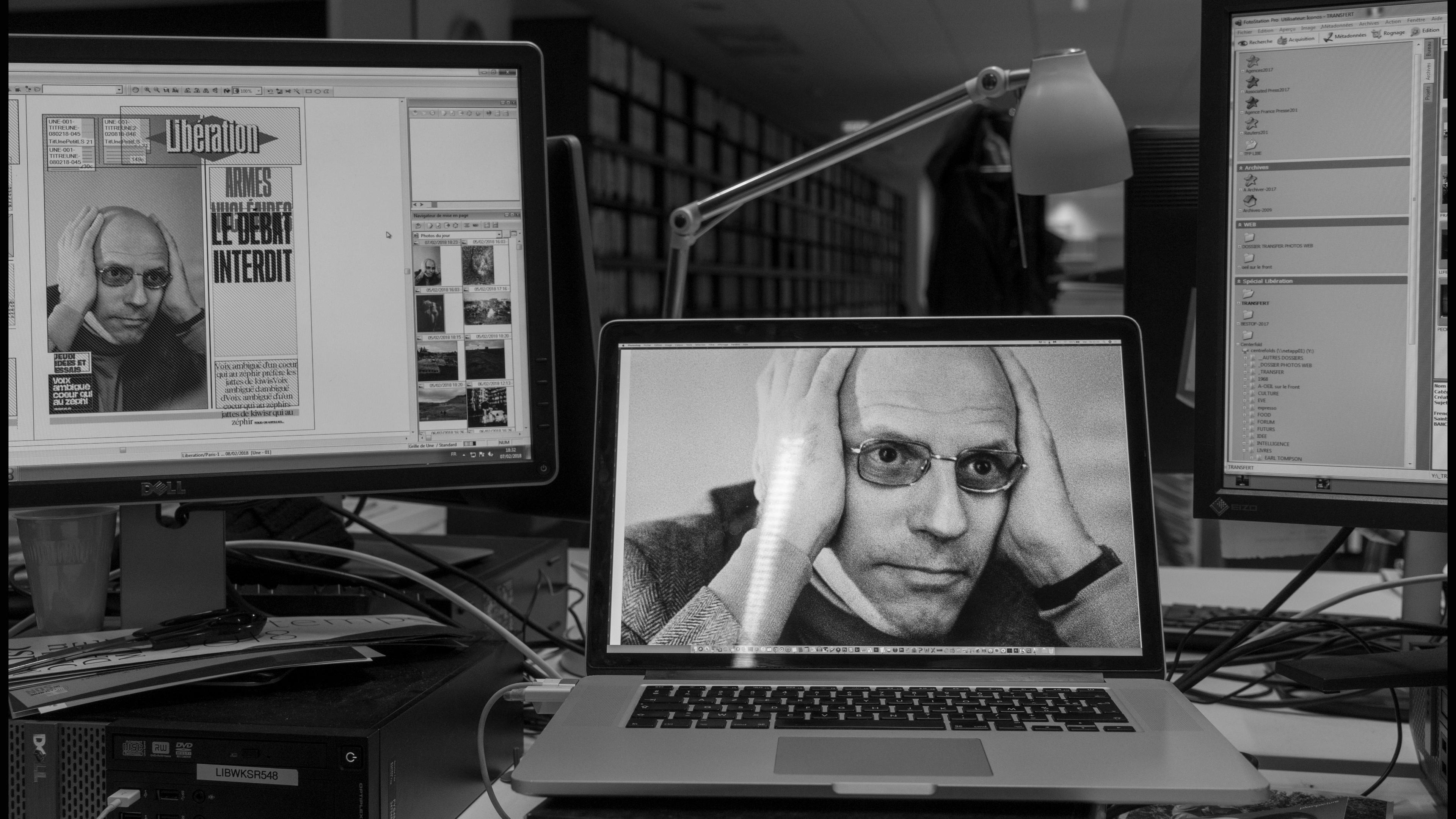 Black and white photo of an office with a laptop and monitors, showing an image of a person with hands on their head.