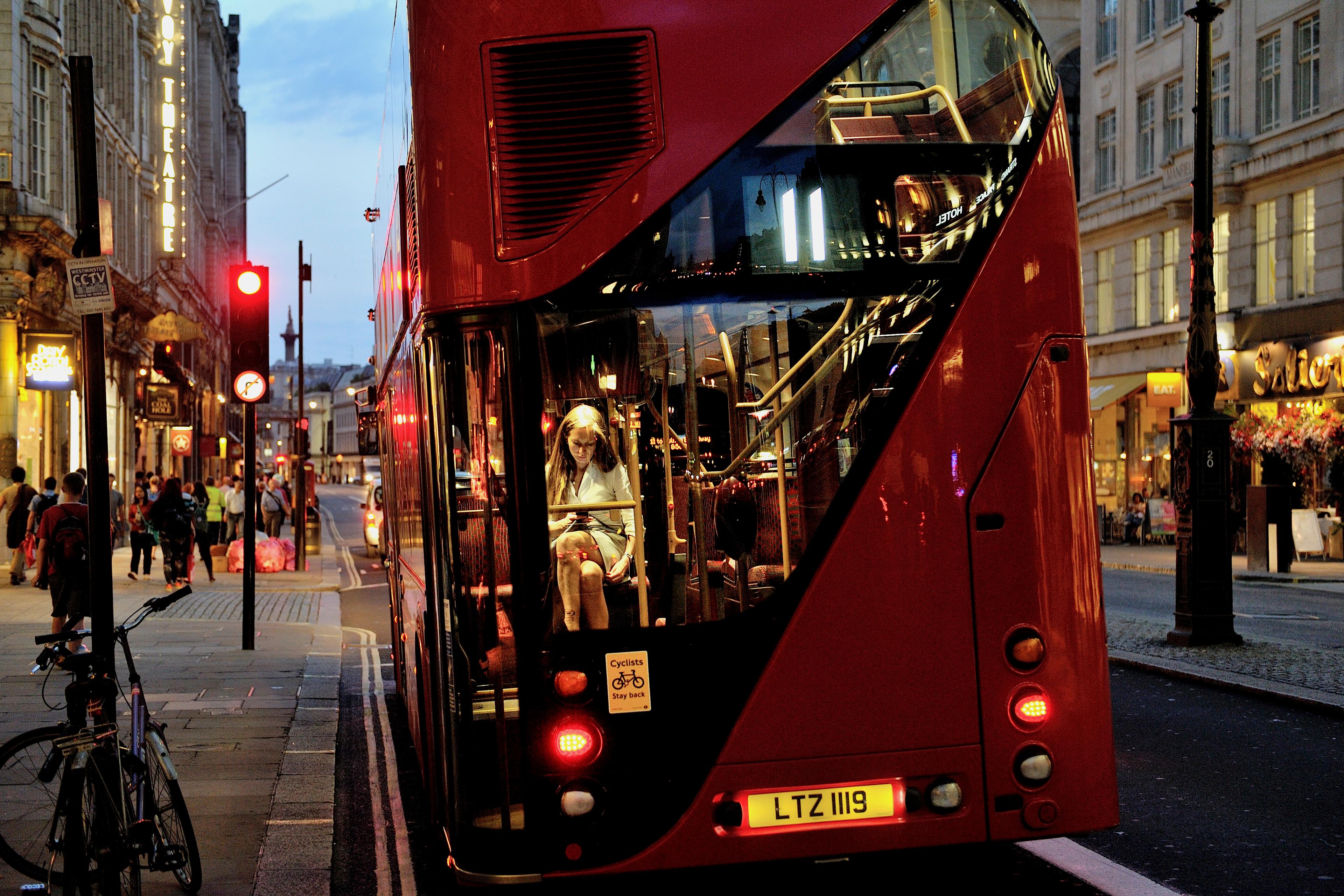 Photo of a red double-decker bus in a city at dusk with a person reading inside pedestrians on the pavement.