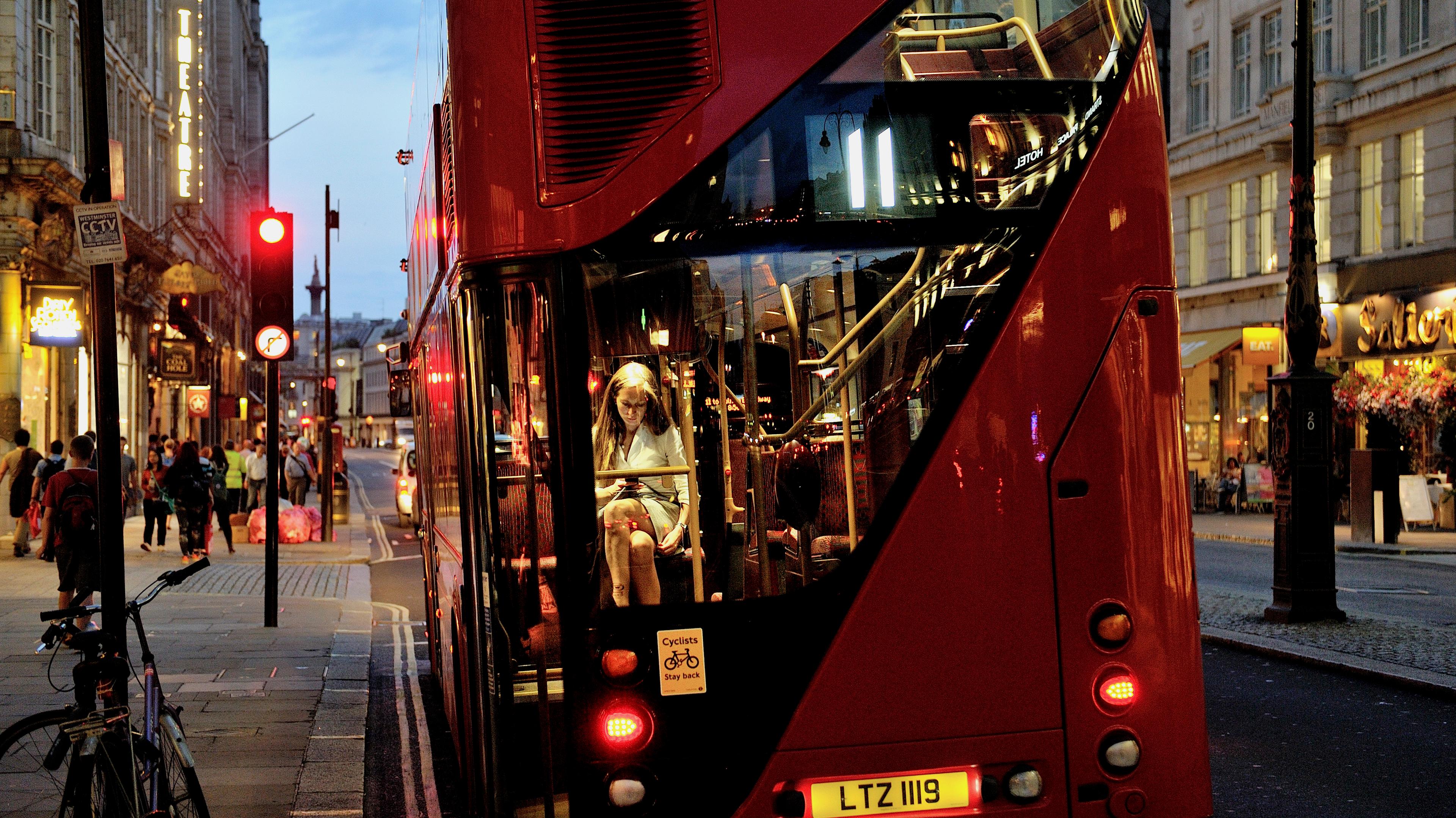 Photo of a red double-decker bus in a city at dusk with a person reading inside pedestrians on the pavement.