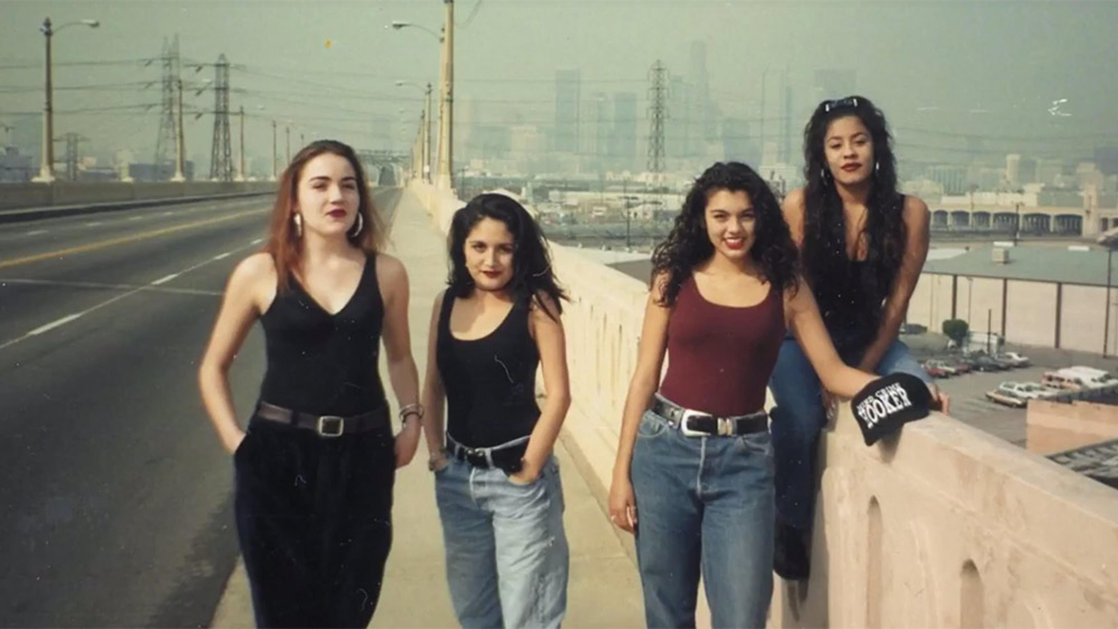 Photo of four young women on a bridge in casual attire, urban background with power lines and buildings, smoggy sky.