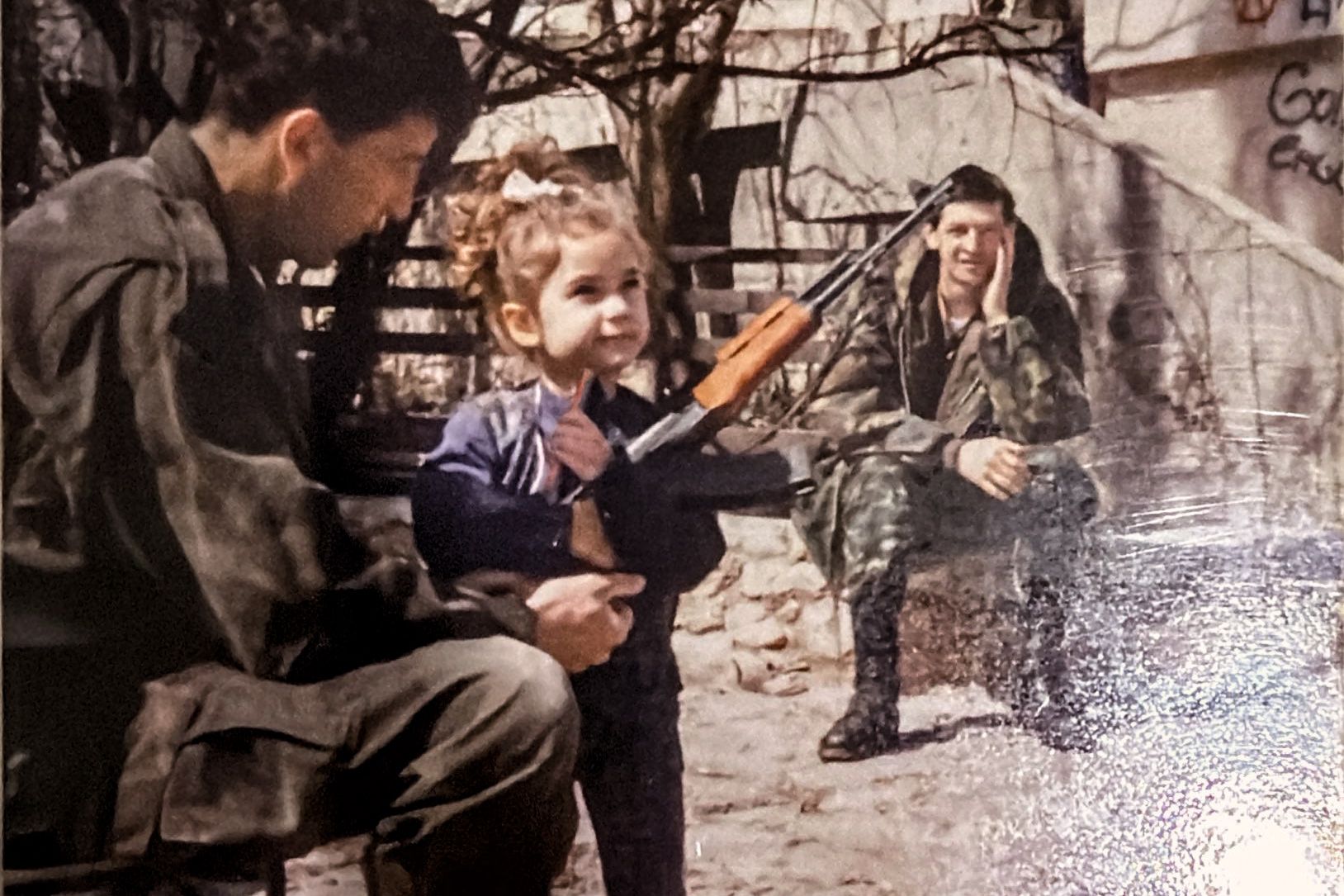 Photo of two soldiers, one helping a child hold a rifle while another soldier sits in the background on a bench.