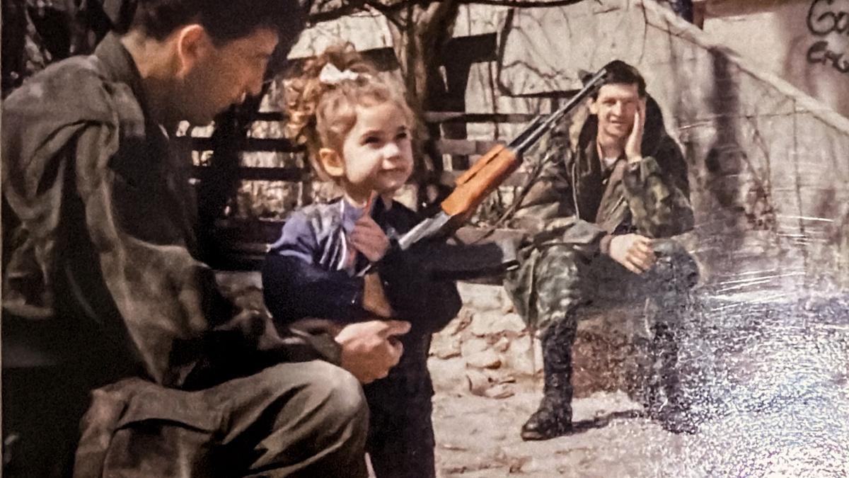 Photo of two soldiers, one helping a child hold a rifle while another soldier sits in the background on a bench.