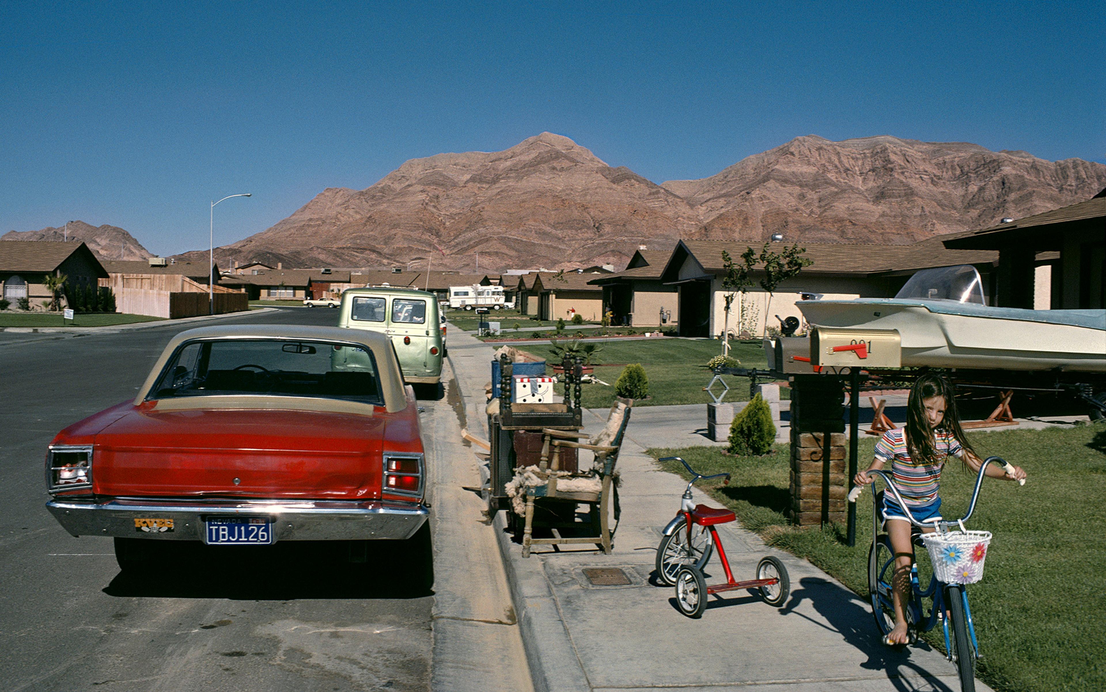 A suburban street with mountains in the background, featuring a girl on a bike, parked cars, and old furniture on the sidewalk in front of a house.