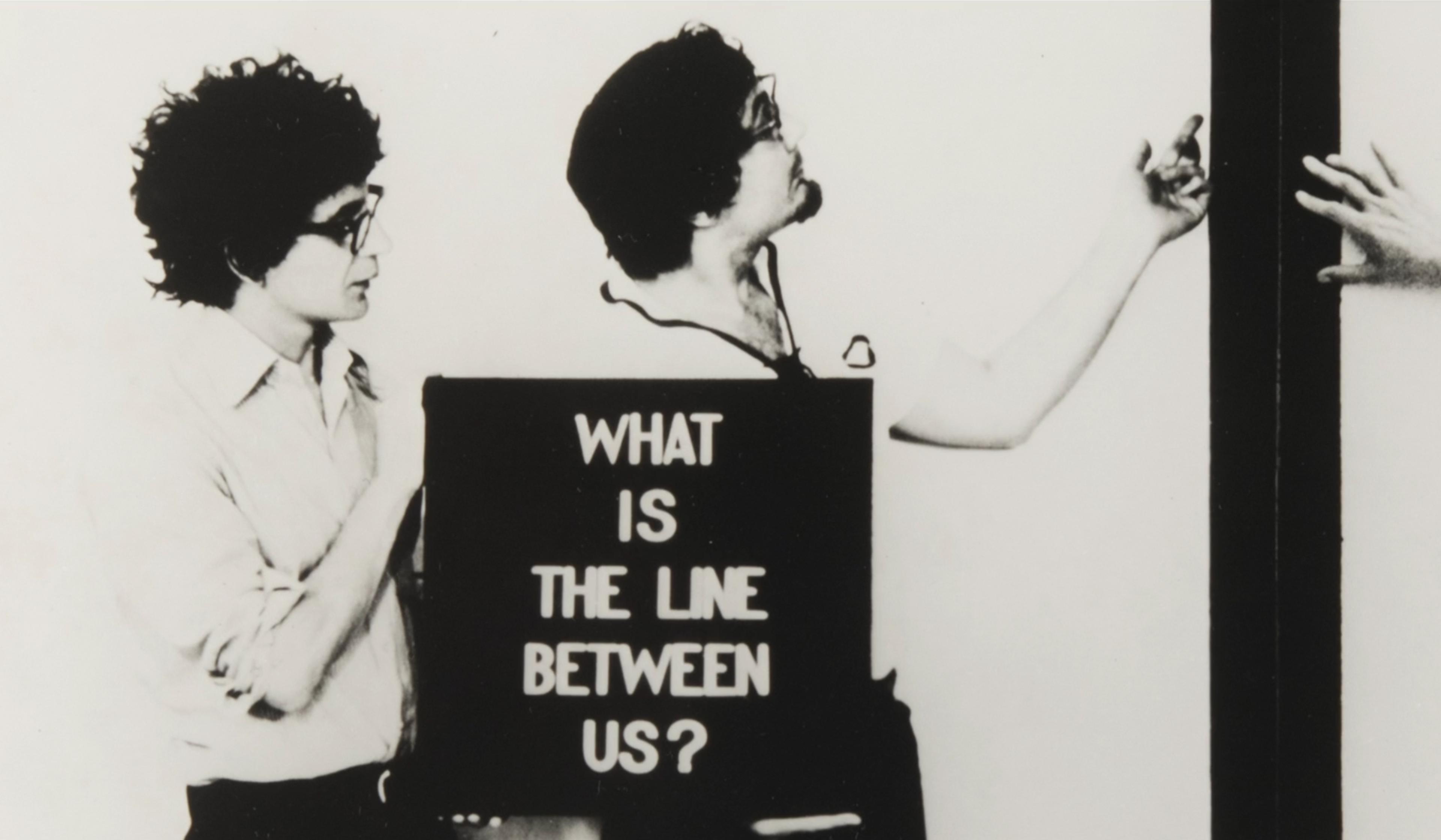 Black-and-white photo of two men standing behind a sign that reads “What is the line between us?”, gesturing towards a partition with another person’s hand visible.