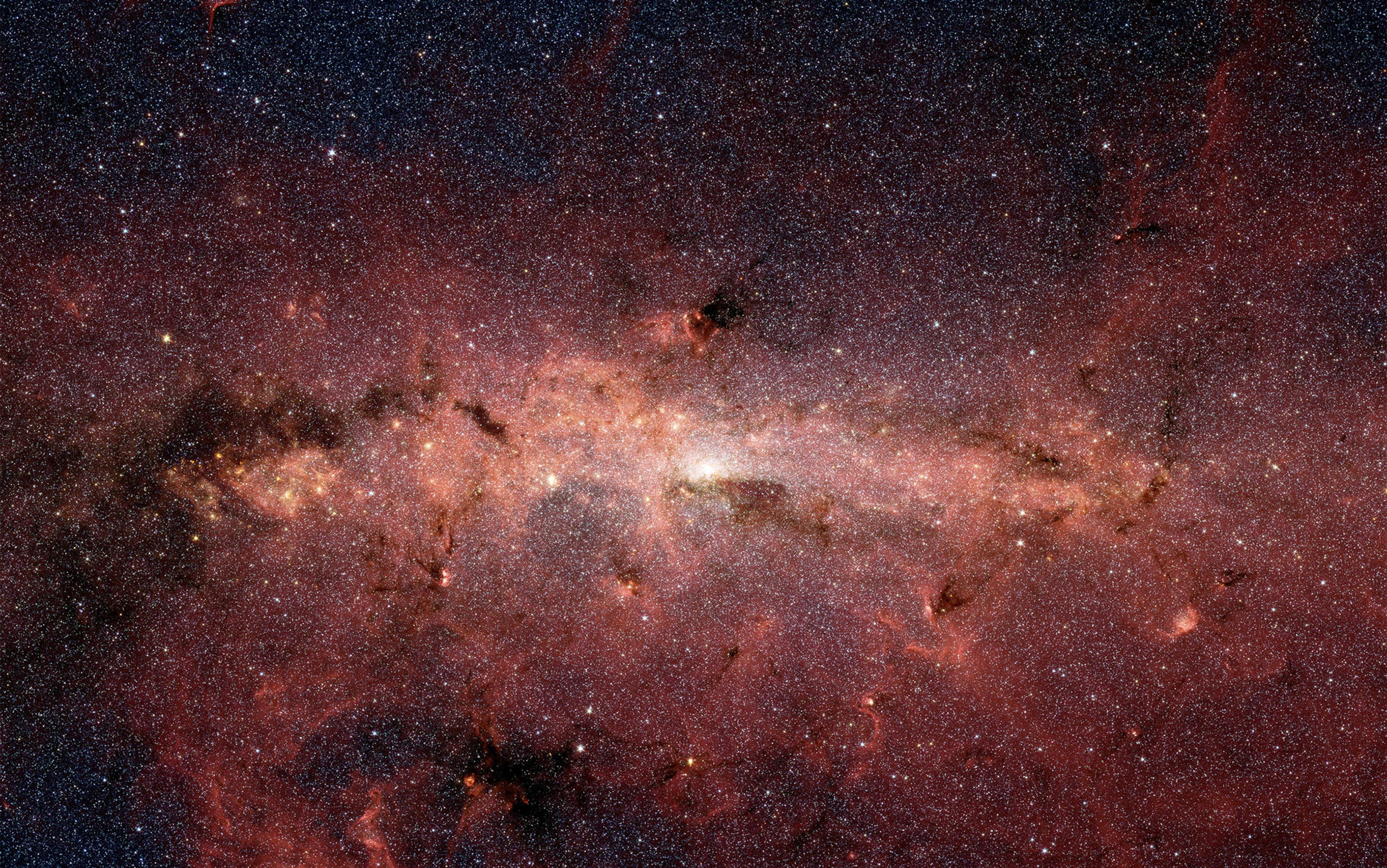 A densely packed star field in the Milky Way’s centre, showing bright stars, dark nebulae, and red, glowing clouds of gas.