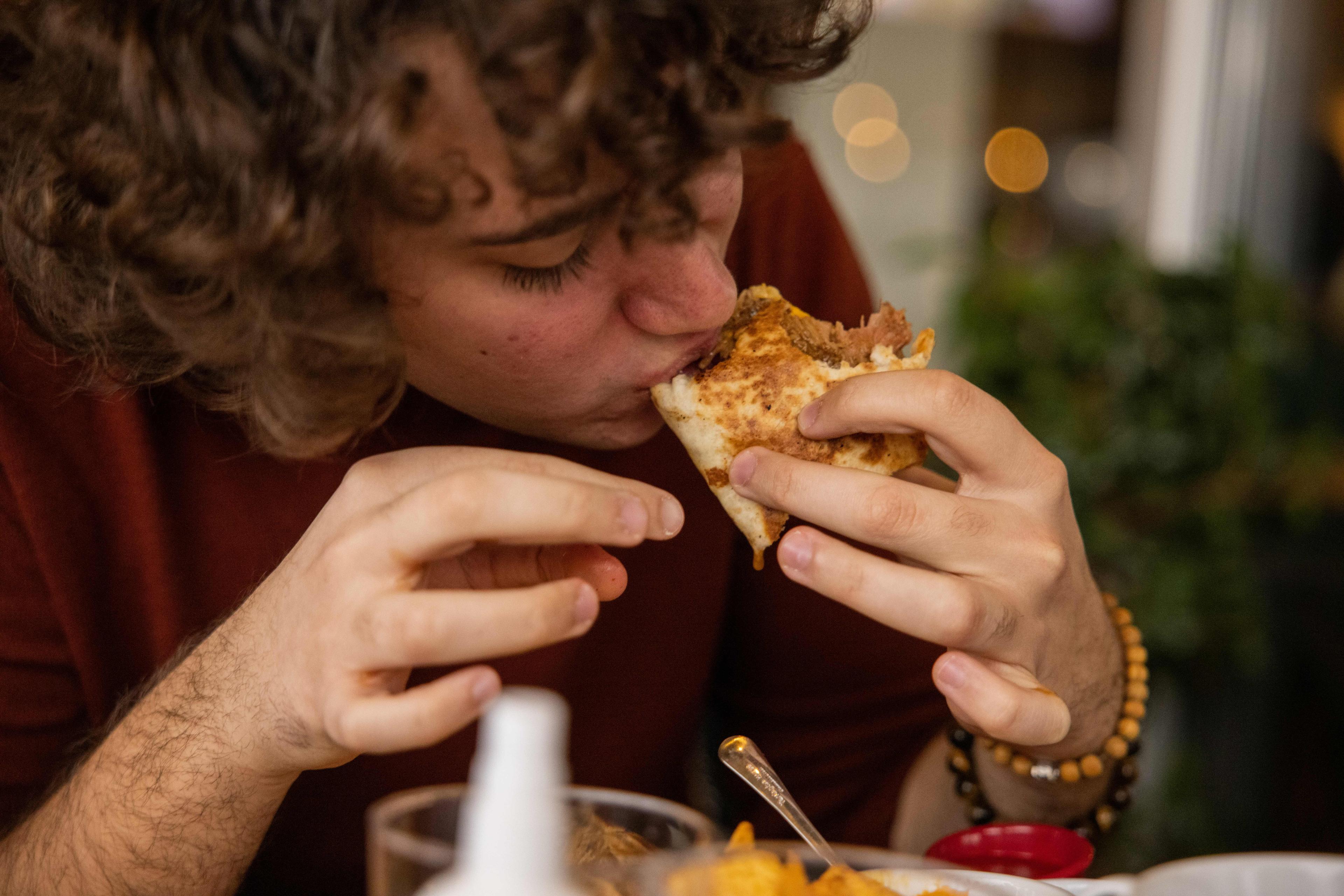 Photo of a person eating a slice of pizza with toppings close-up, in a cosy indoor setting with bokeh lights in background.