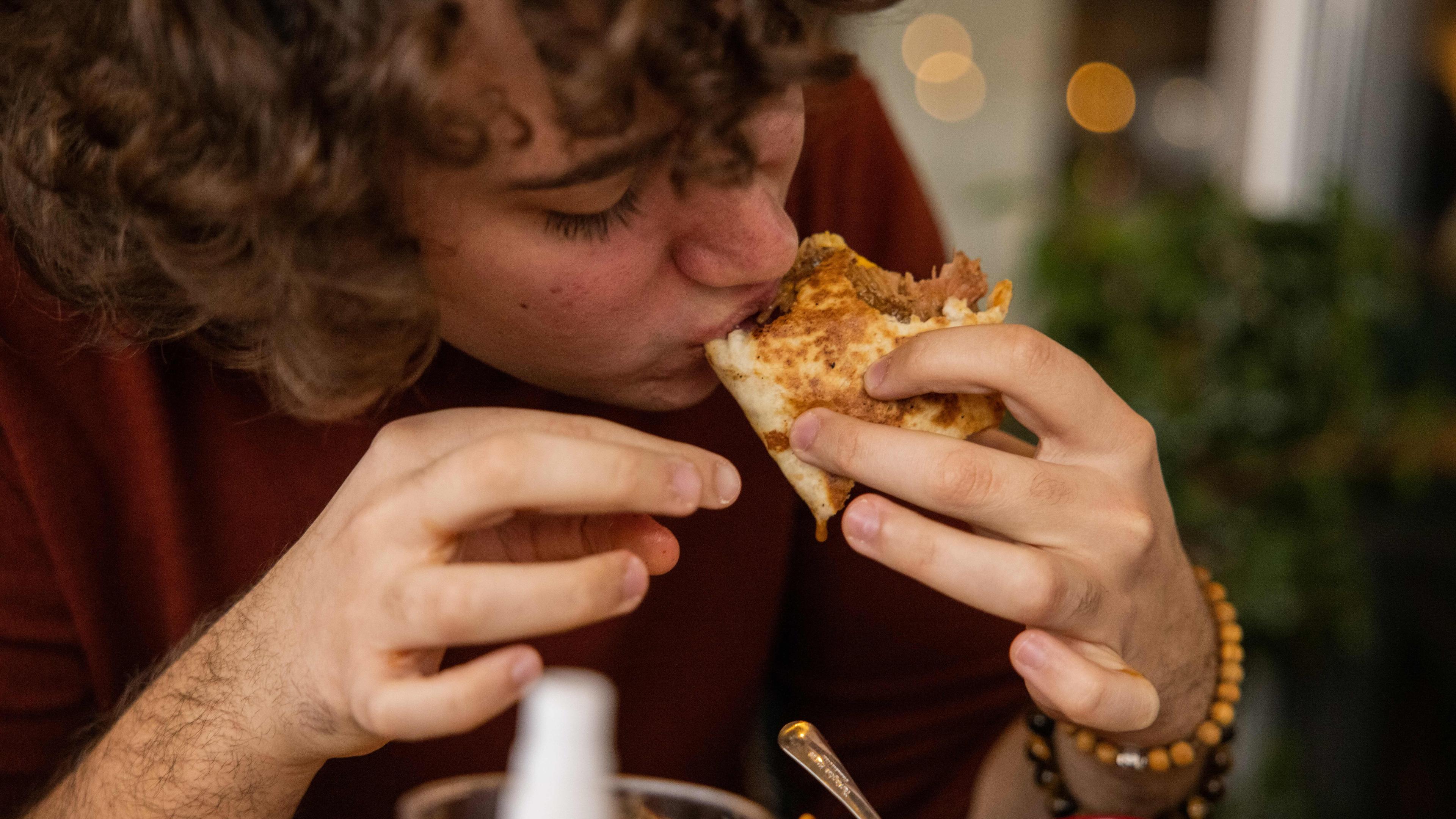 Photo of a person eating a slice of pizza with toppings close-up, in a cosy indoor setting with bokeh lights in background.