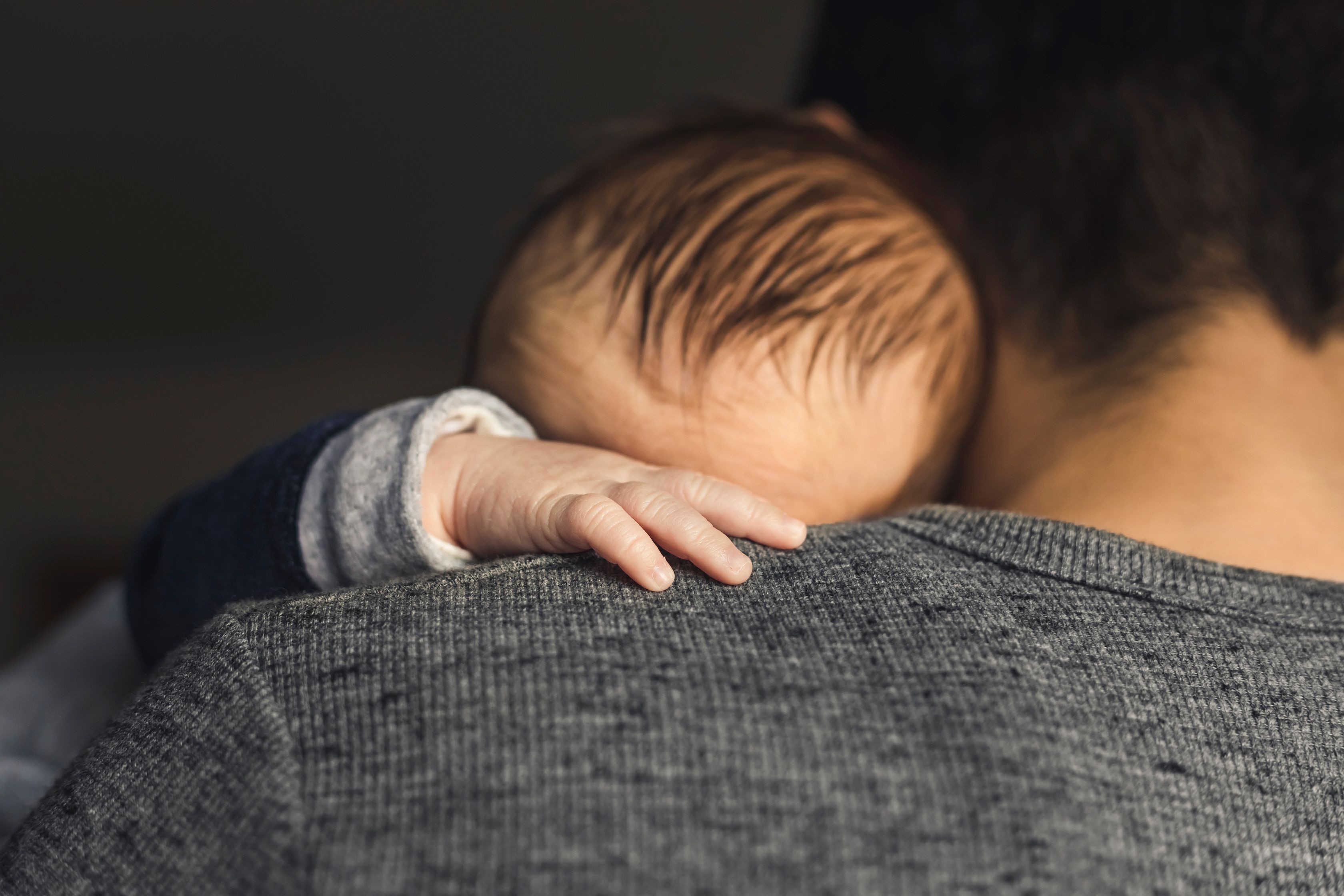 A baby resting on an adult’s shoulder, with the baby’s hand visible on a grey jumper against a dark background.