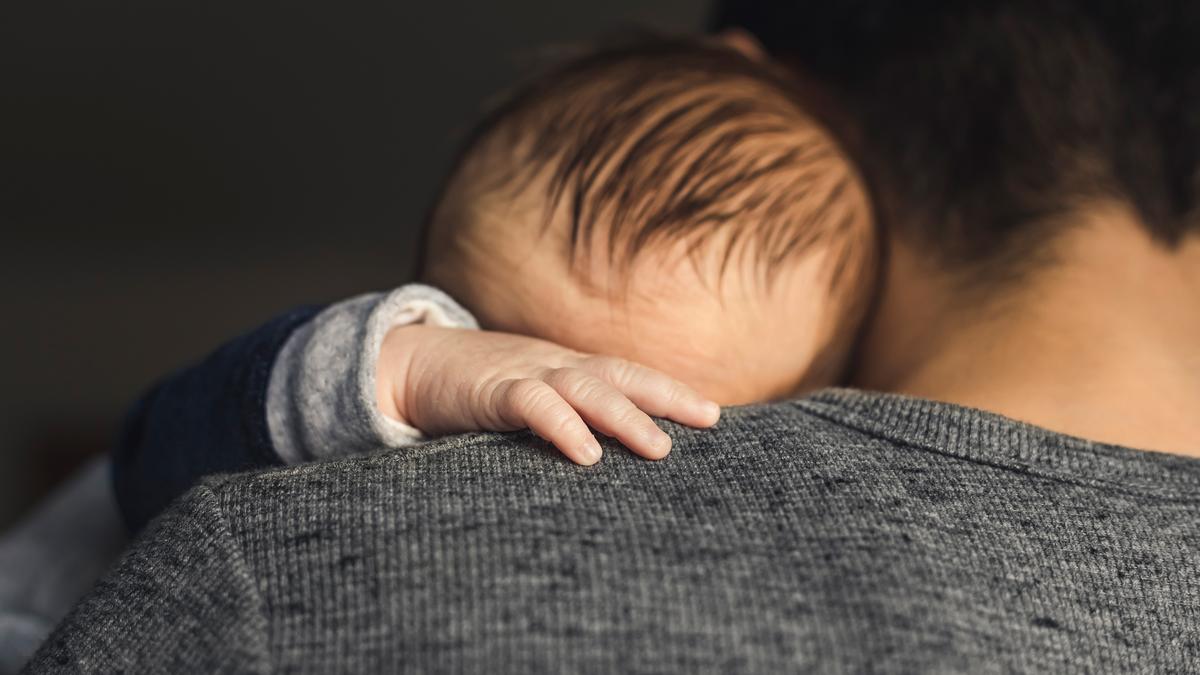 A baby resting on an adult’s shoulder, with the baby’s hand visible on a grey jumper against a dark background.