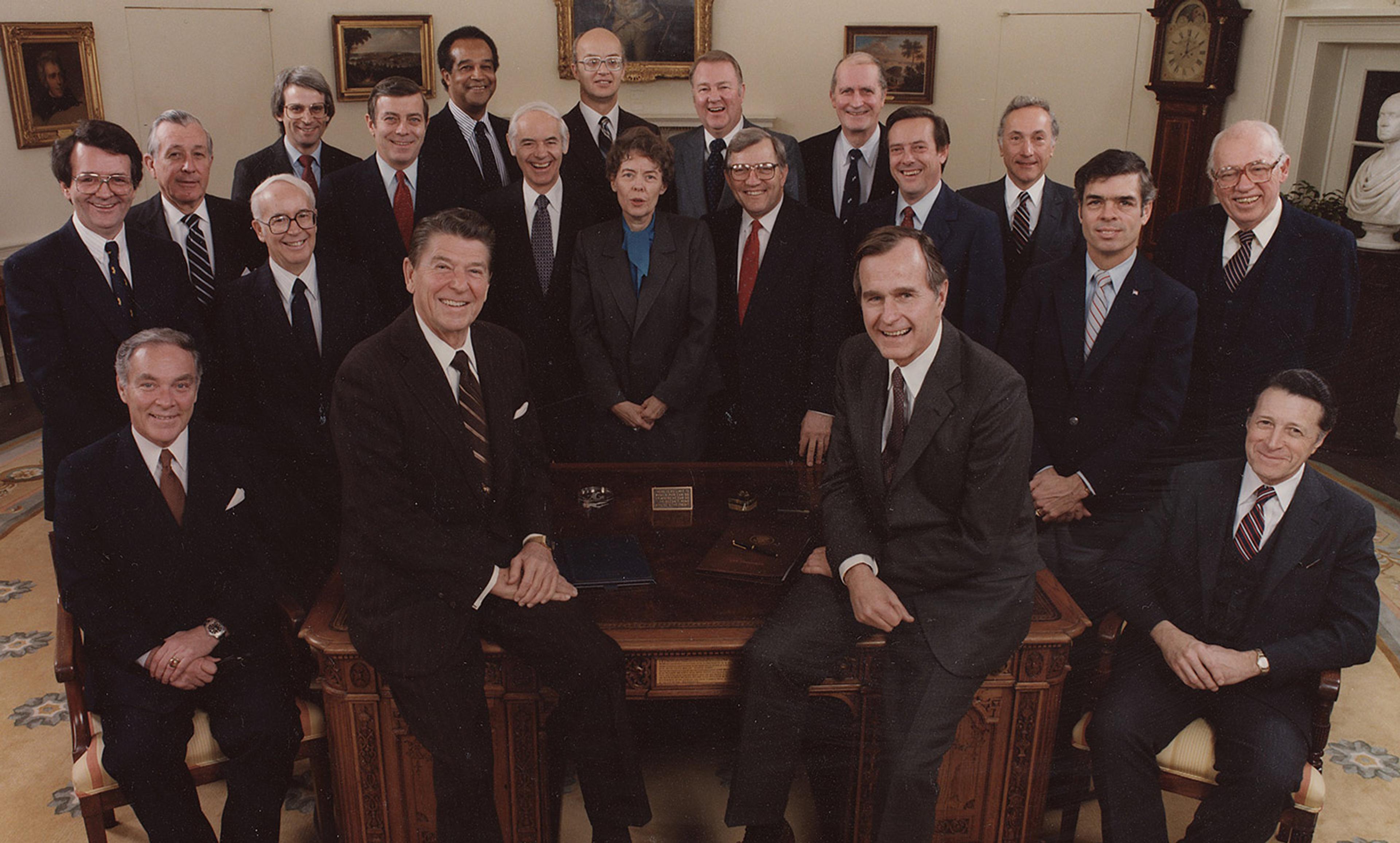 A group of 21 men, including Ronald Reagan and George Bush Sr, and one woman in formal attire, posed in an office with paintings and a grandfather clock.