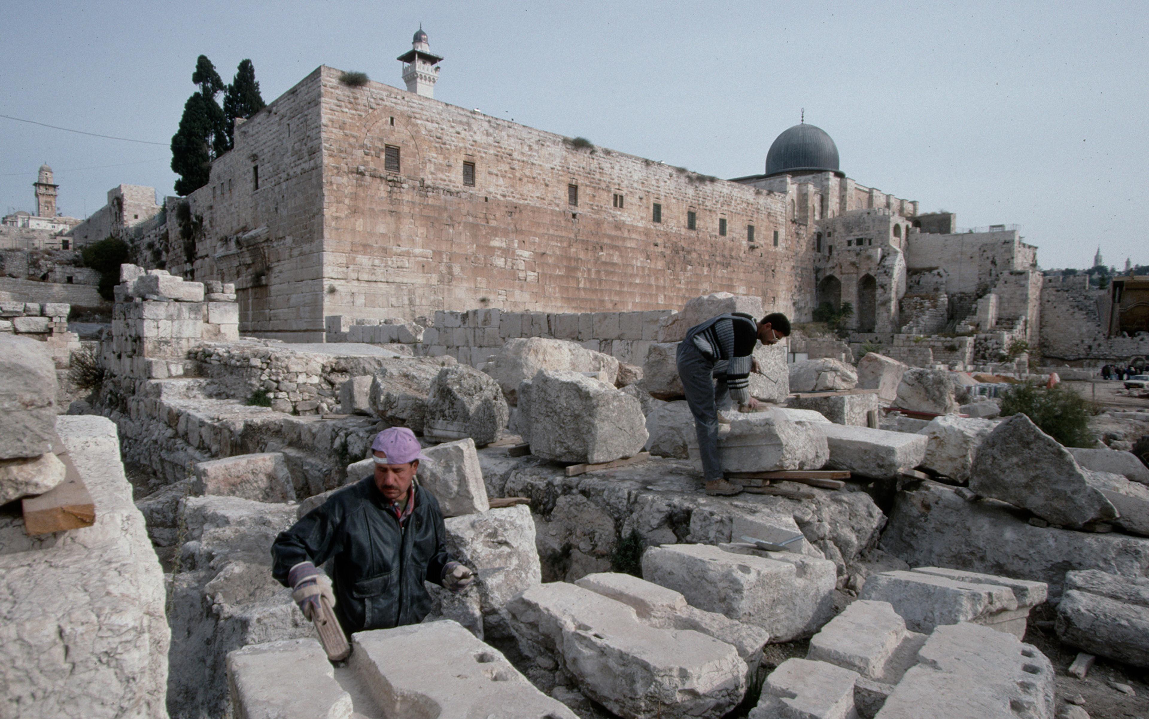 Two men dressed in work attire examining and restoring ancient stone ruins in front of a historic stone building with a domed roof.
