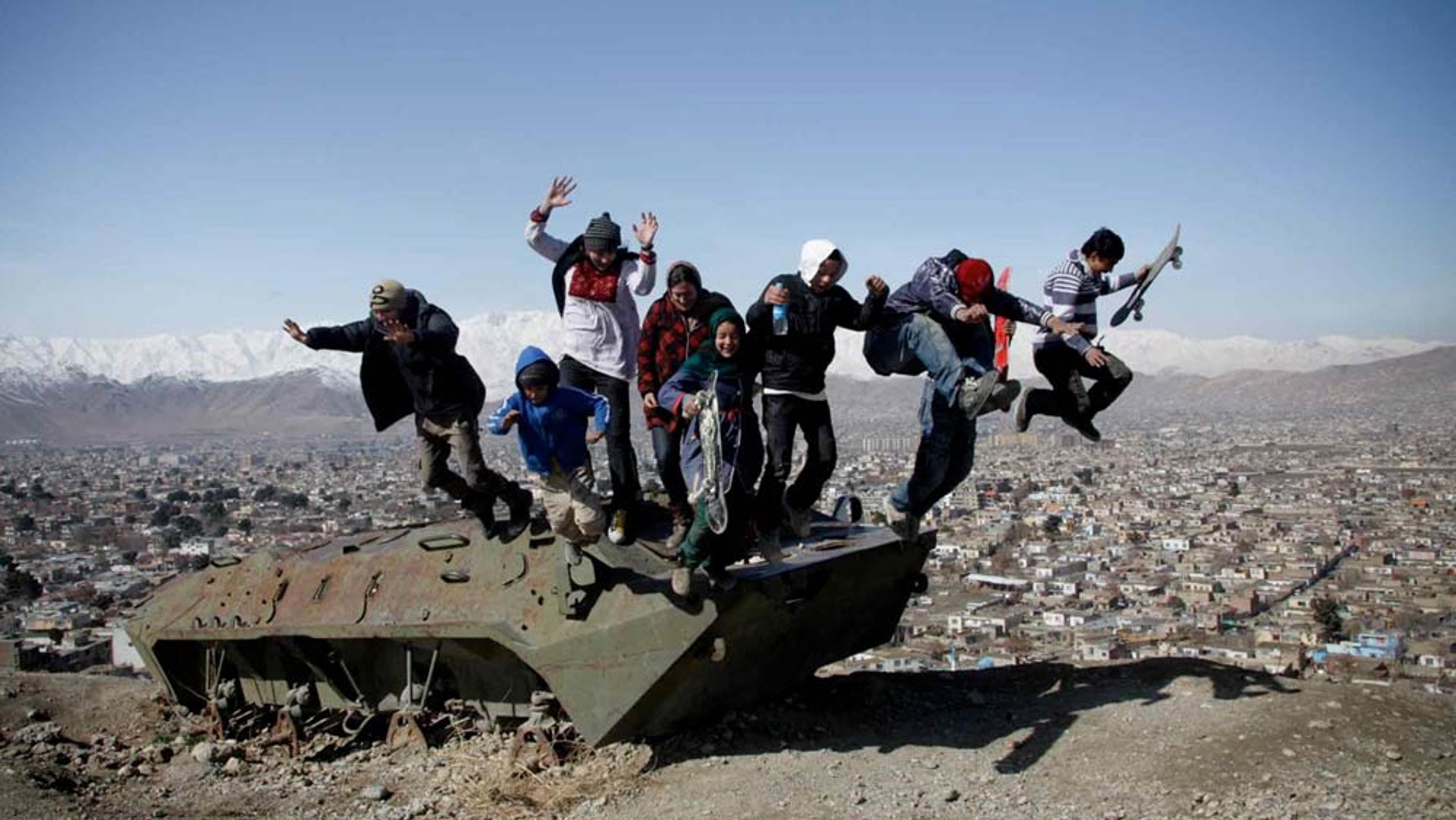 Children jumping off an old tank overlooking a city with snowy mountains in the background on a sunny day.