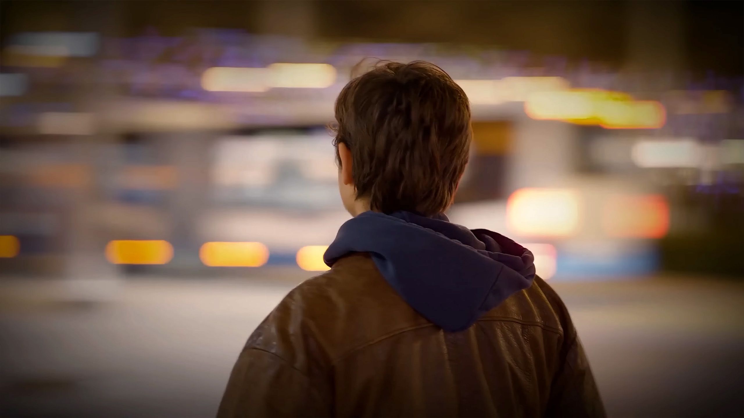 Photo of a person with short hair wearing a brown jacket and blue hoodie, facing blurry city lights at night.