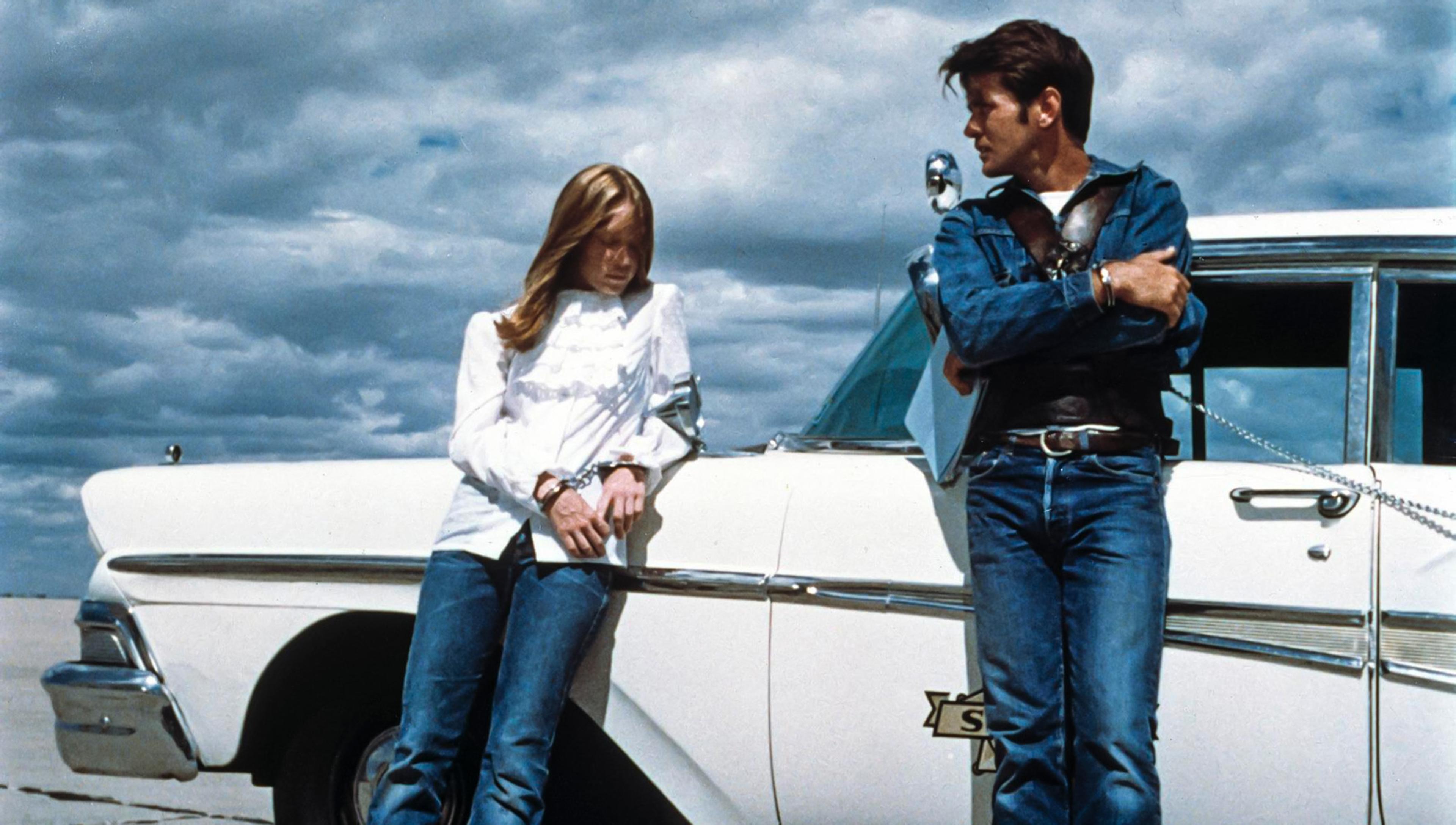 A young couple in handcuffs lean against a 1950s police car beneath a brooding prairie sky