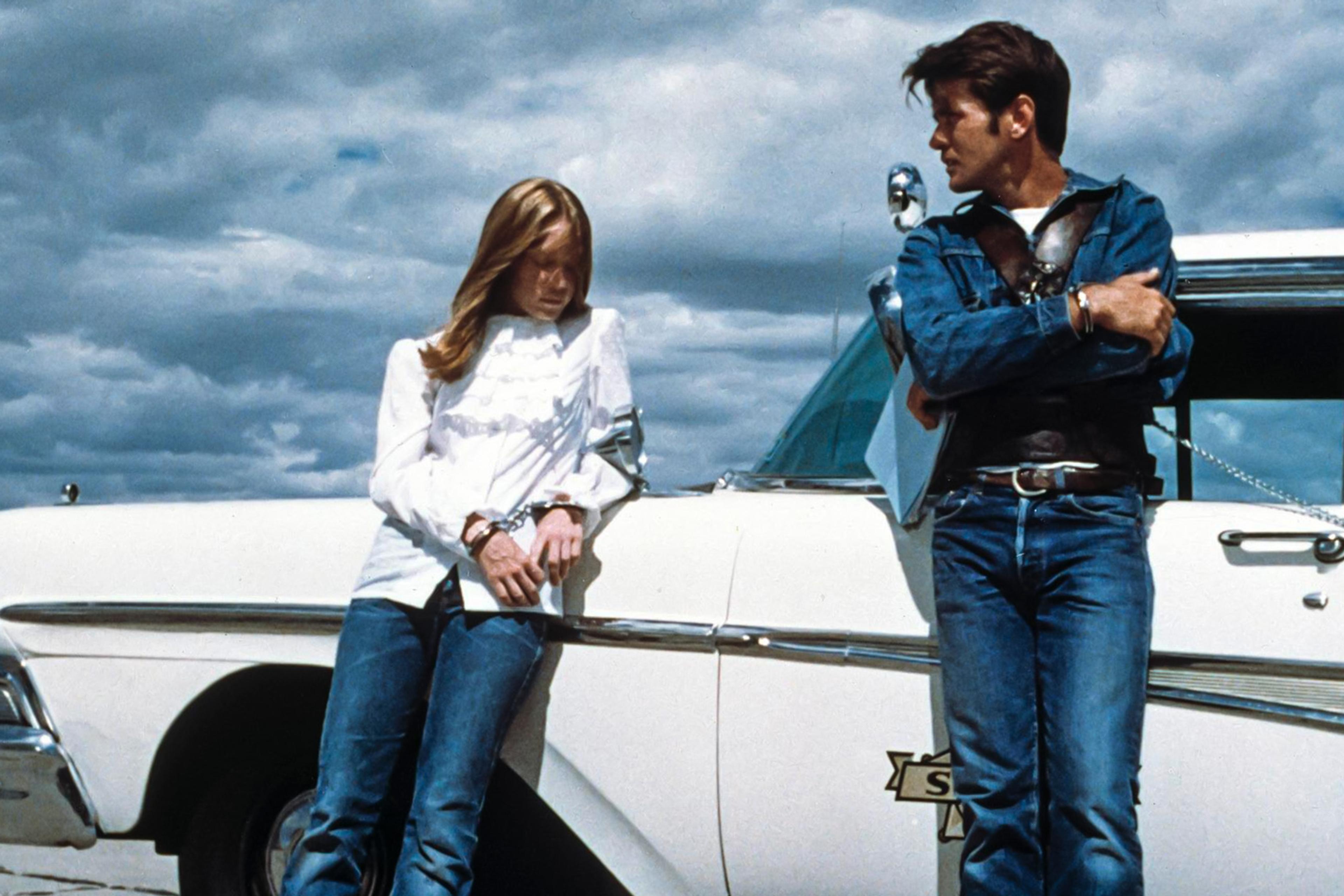 A young couple in handcuffs lean against a 1950s police car beneath a brooding prairie sky
