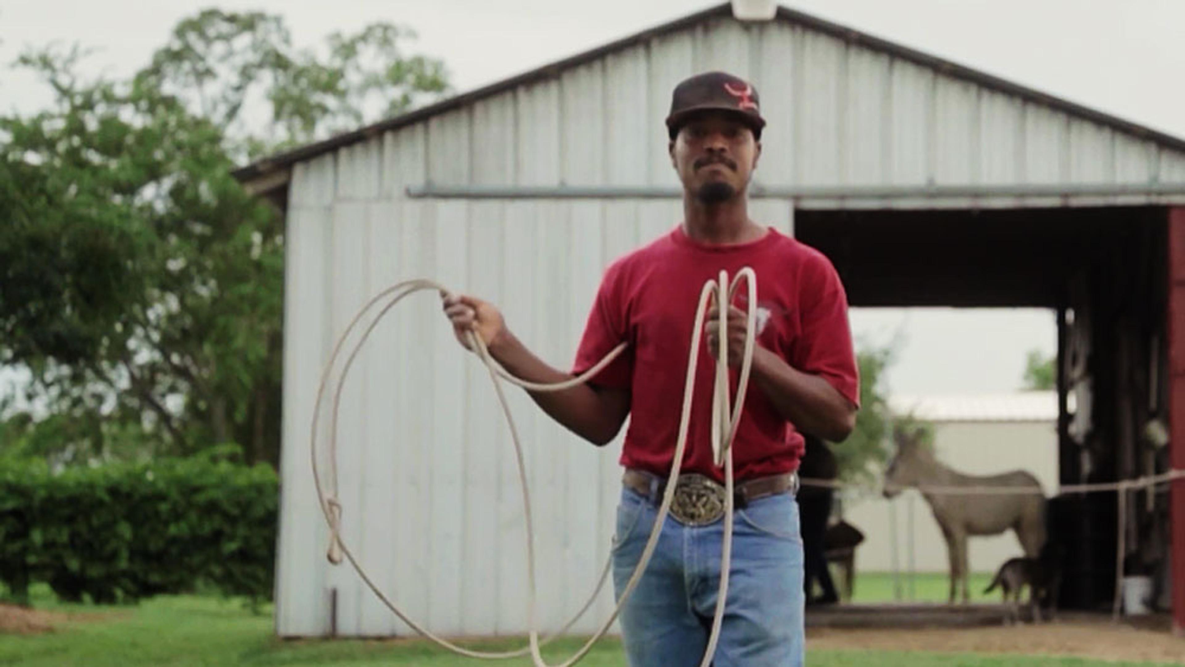 A man in a red shirt holding a rope in front of a barn with trees in the background and a horse inside the barn.