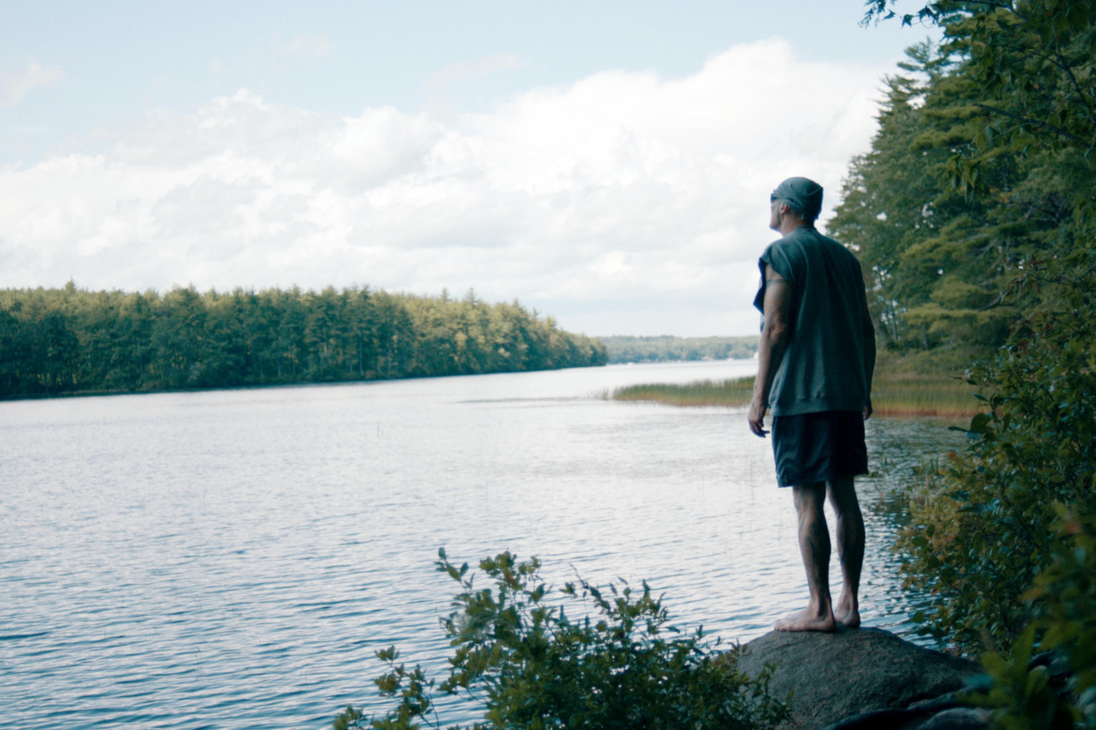 A man recently released from prison standing on a rock by a tranquil lake, surrounded by dense green trees and under a partly cloudy sky.