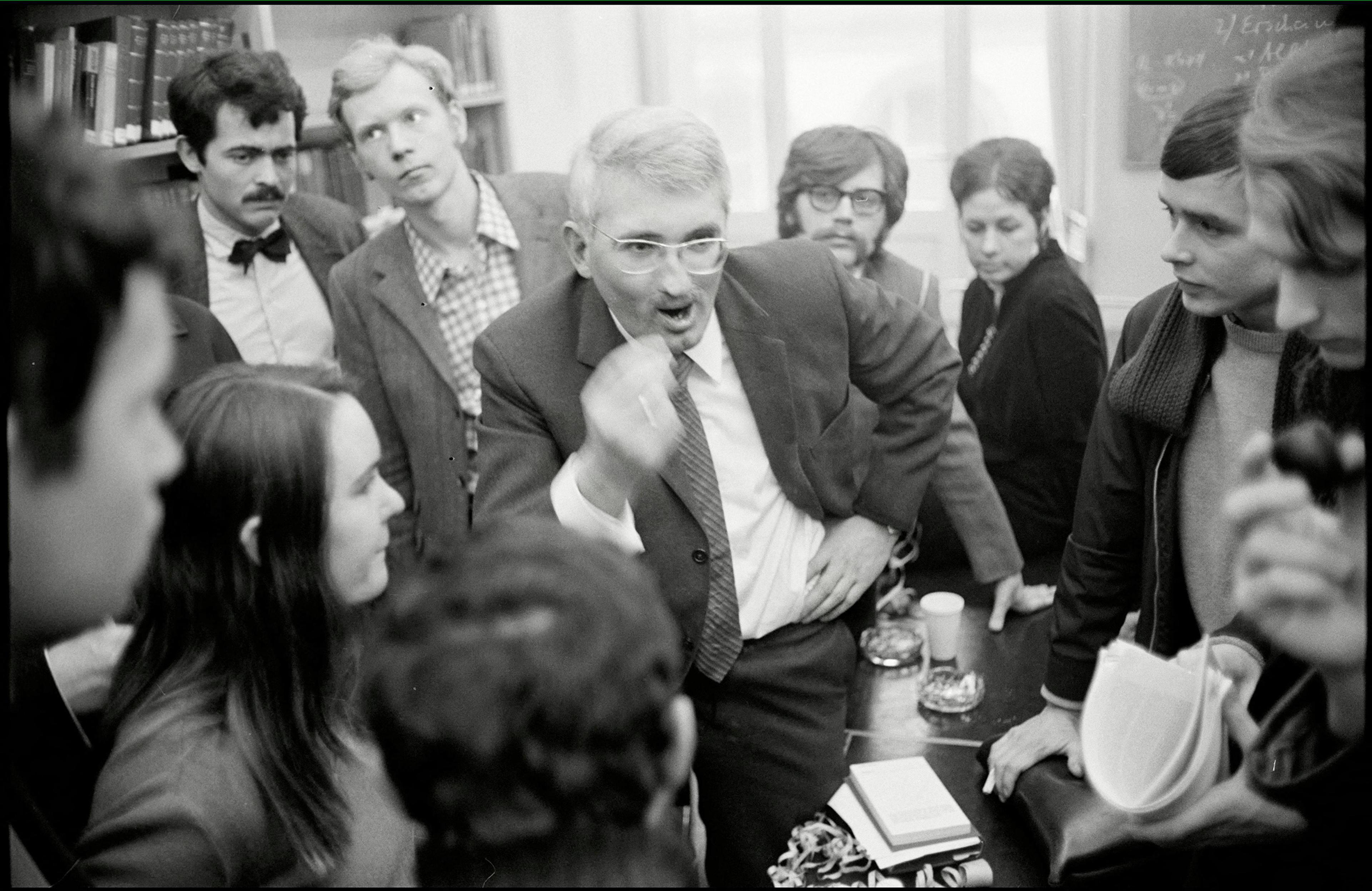 Black and white photo of a man in glasses animatedly speaking to a group gathered around him in an indoor setting.