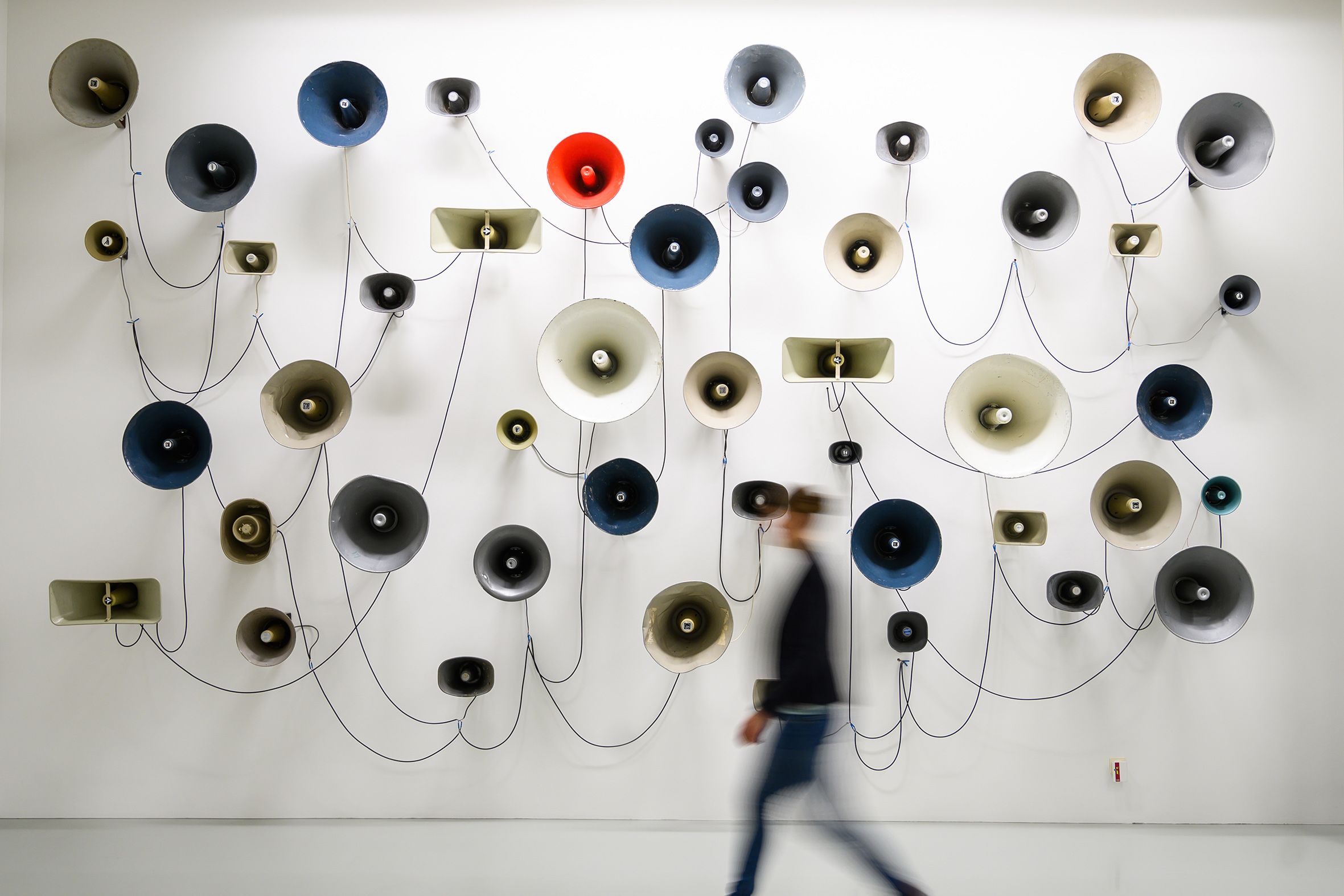Art installation photo of various coloured megaphones on a wall with a blurred person walking by.