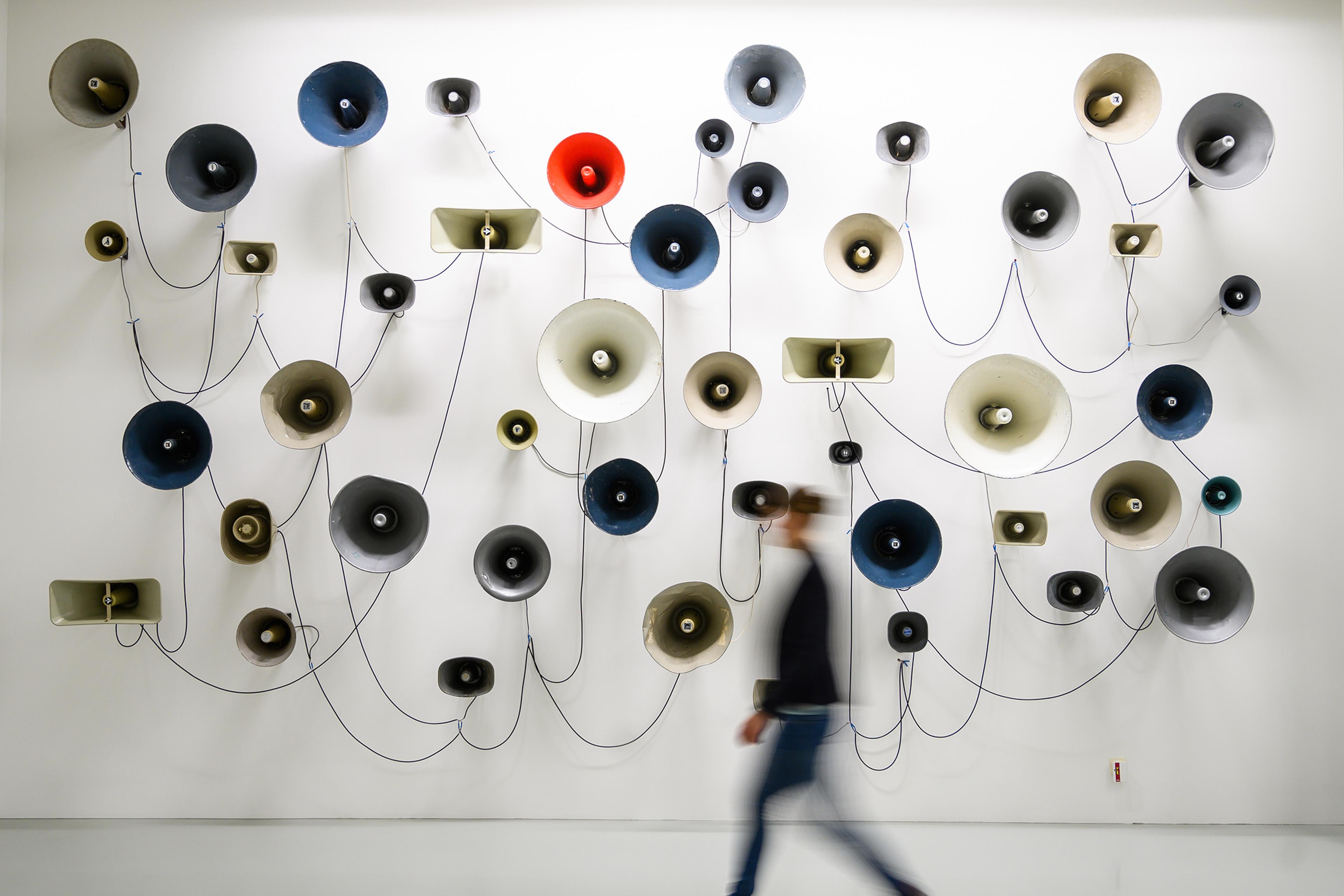 Art installation photo of various coloured megaphones on a wall with a blurred person walking by.
