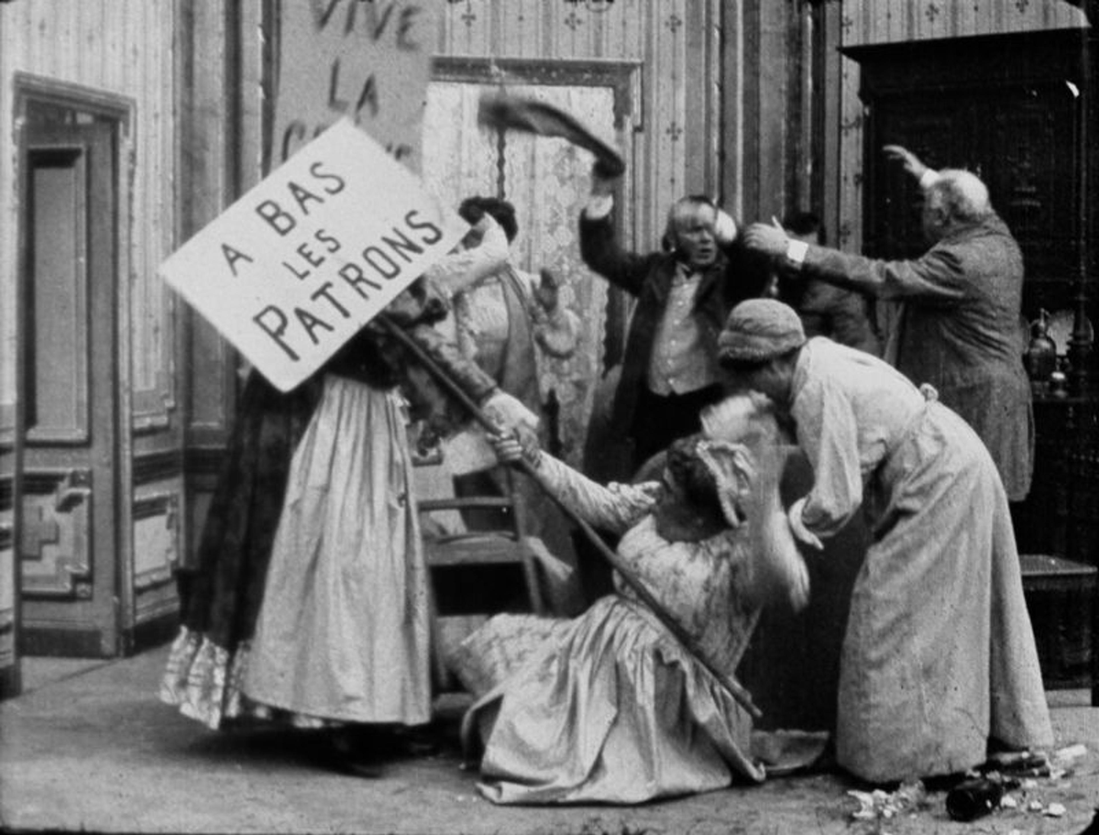 Black-and-white photo of woman in period clothing engaged in a chaotic scene with protest signs in a room with vintage decor.