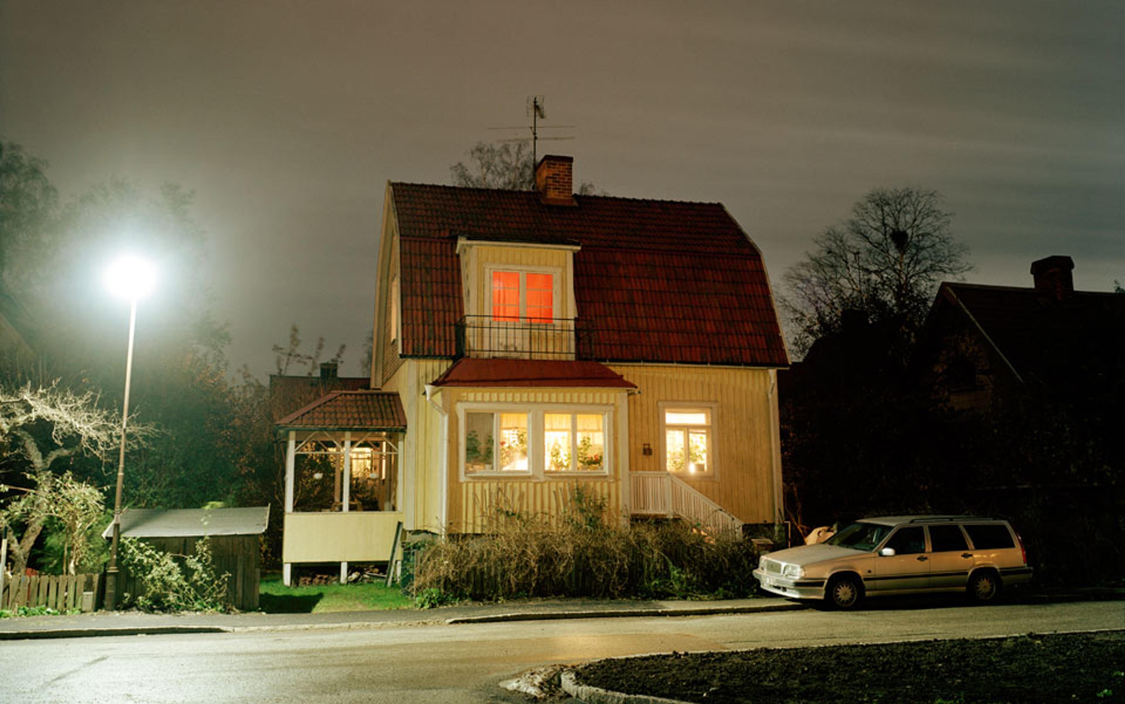 A two-storey house at night with lights on inside, a car parked outside, and a streetlamp illuminating the scene.