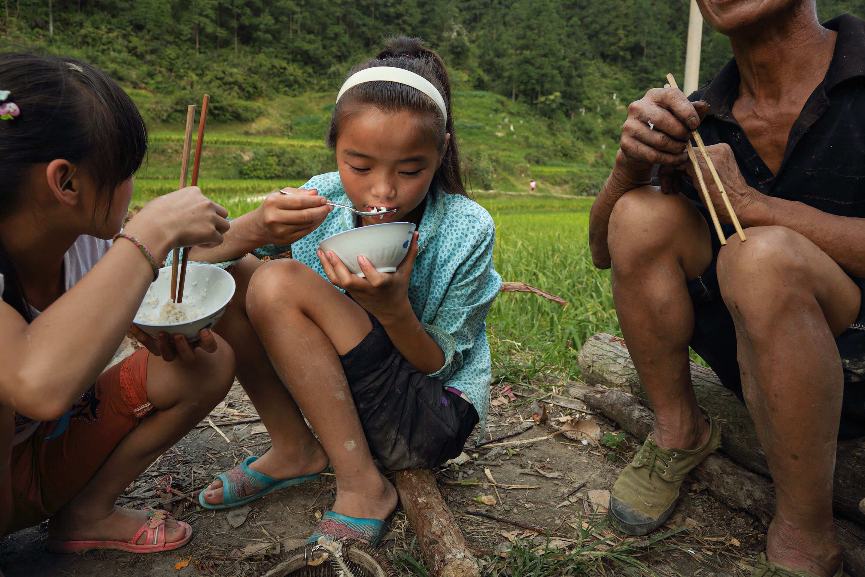 Children sitting outdoors eating from bowls with chopsticks or spoons next to a seated adult in a green field.
