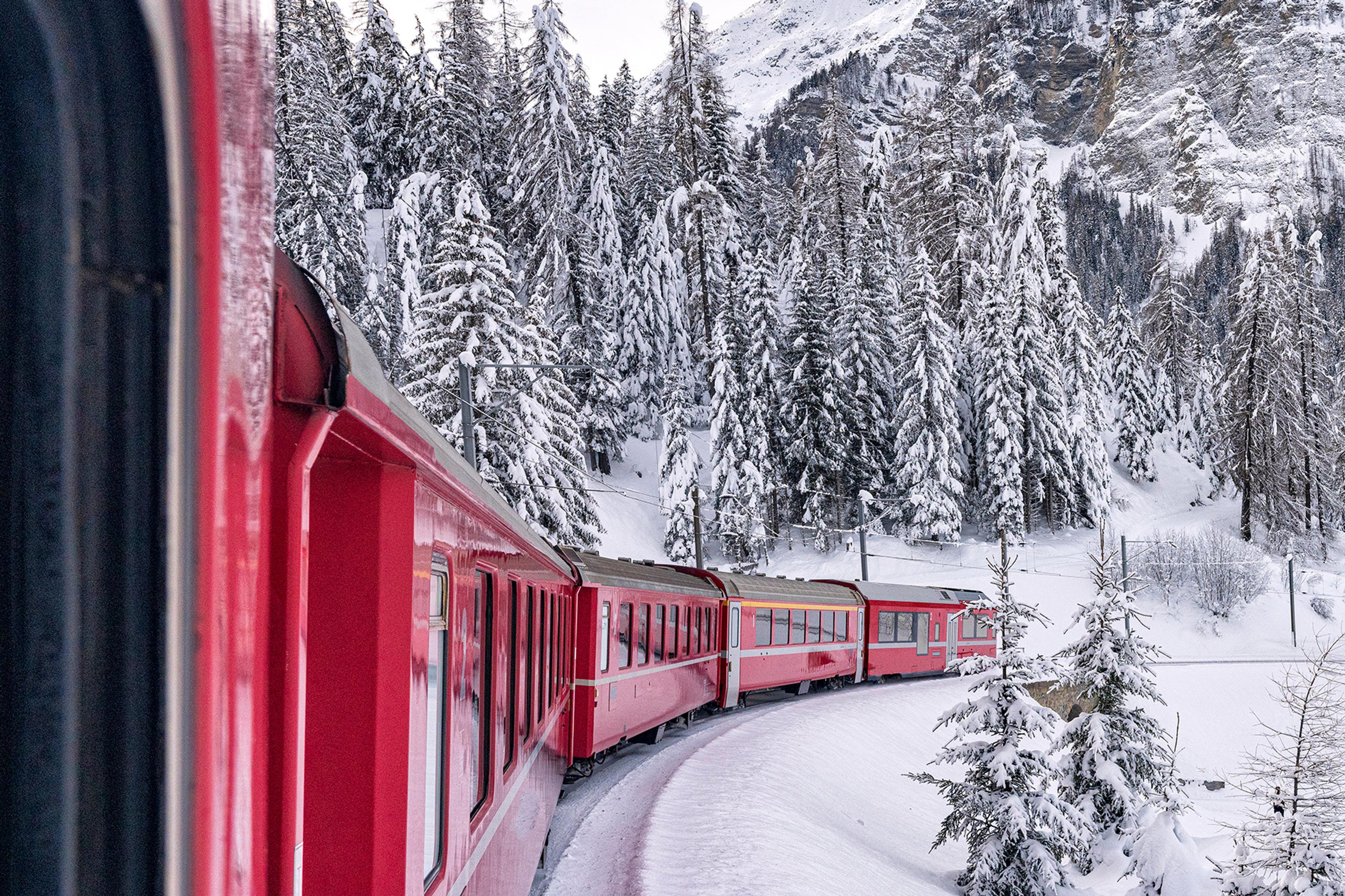 Red train travelling through a snow-covered mountainous landscape with frosted pine trees and snowy hills, viewed from inside one of the carriages.