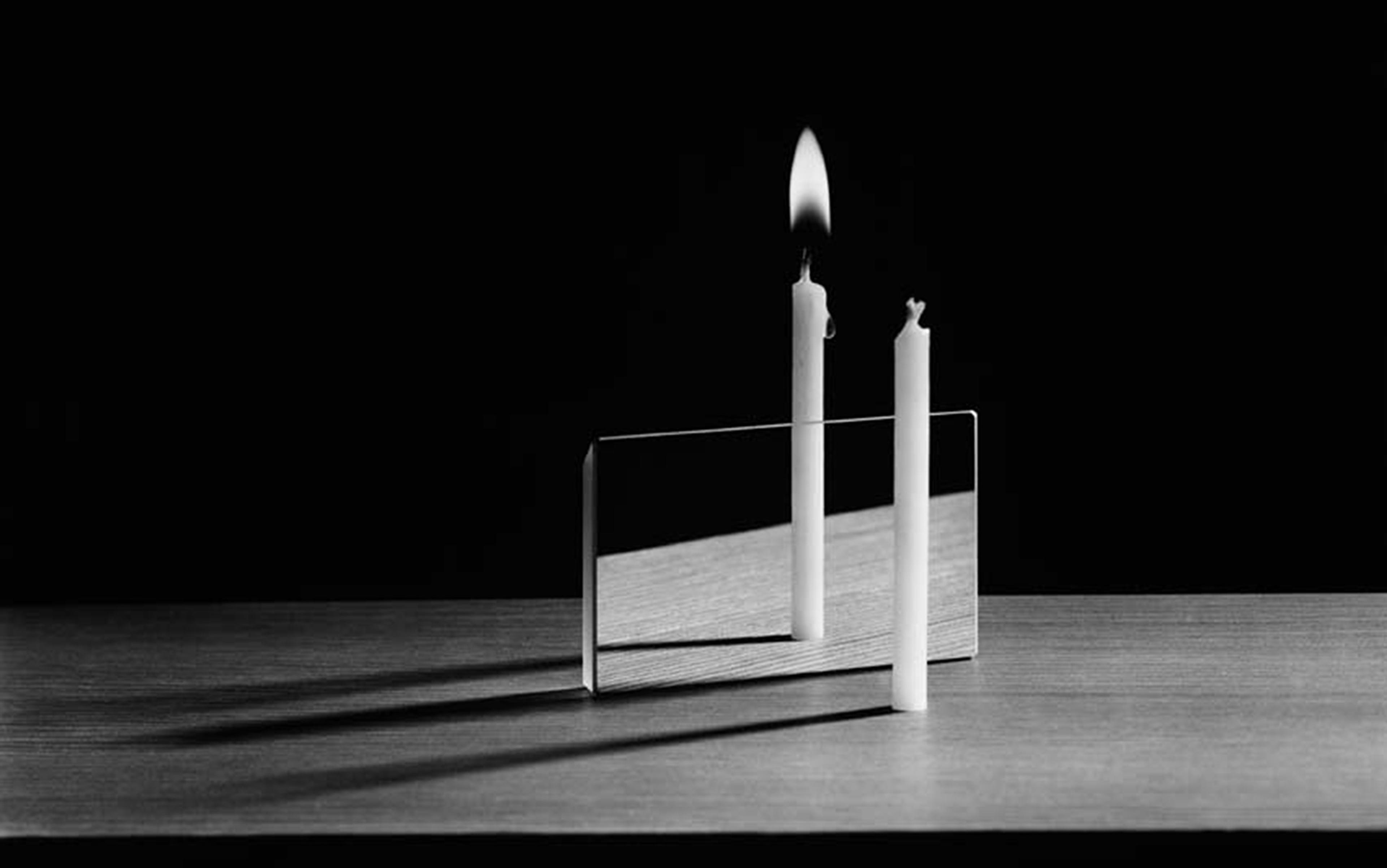 An unlit candle beside a lit one reflected in a mirror creating the illusion both are lit, on a table with a dark background.