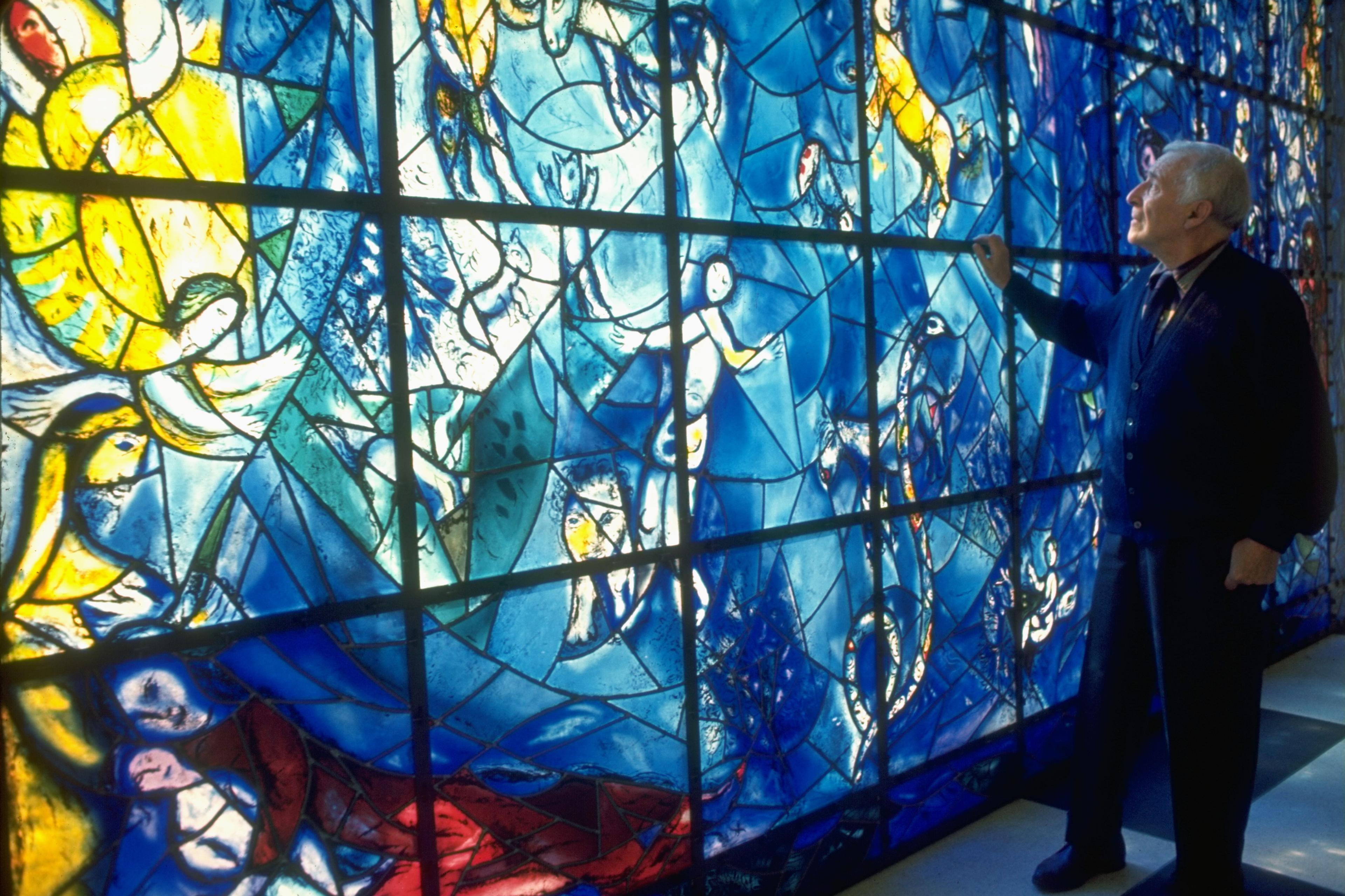 Photo of a man admiring a large stained glass artwork with vibrant blue and yellow hues depicting various figures and animals.