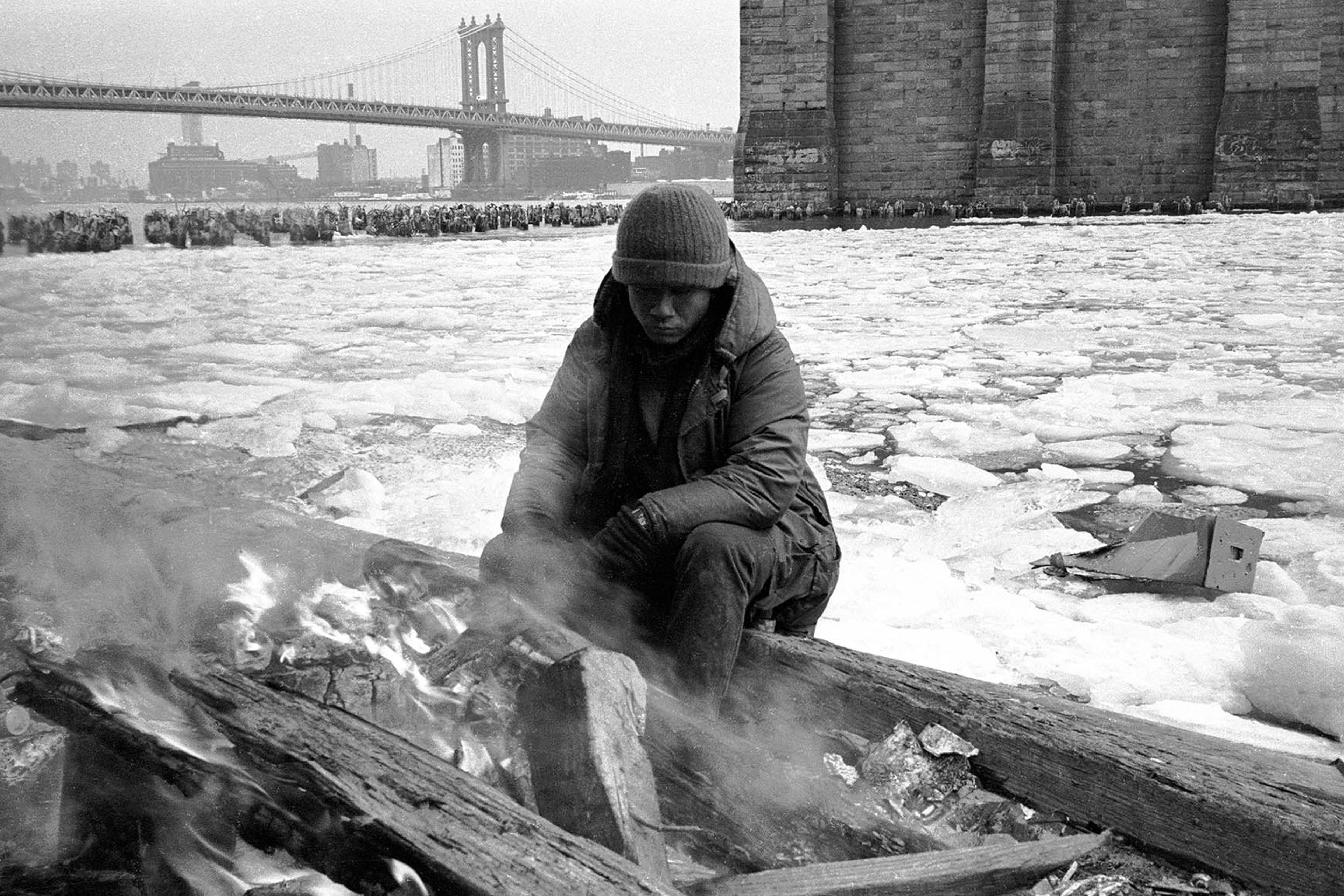 Black-and-white photo: person sitting by a fire on a snowy riverbank under a bridge, wearing a winter coat and hat.