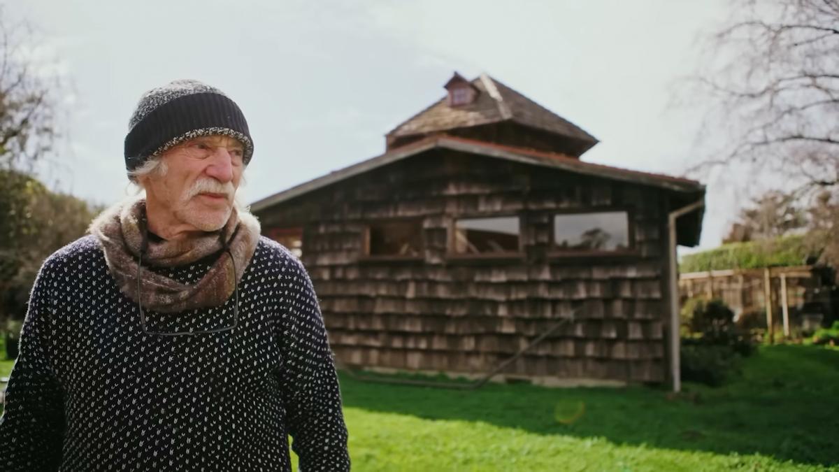 Photo of an elderly man in a hat and scarf standing in a grassy yard with a rustic wooden building in the background.