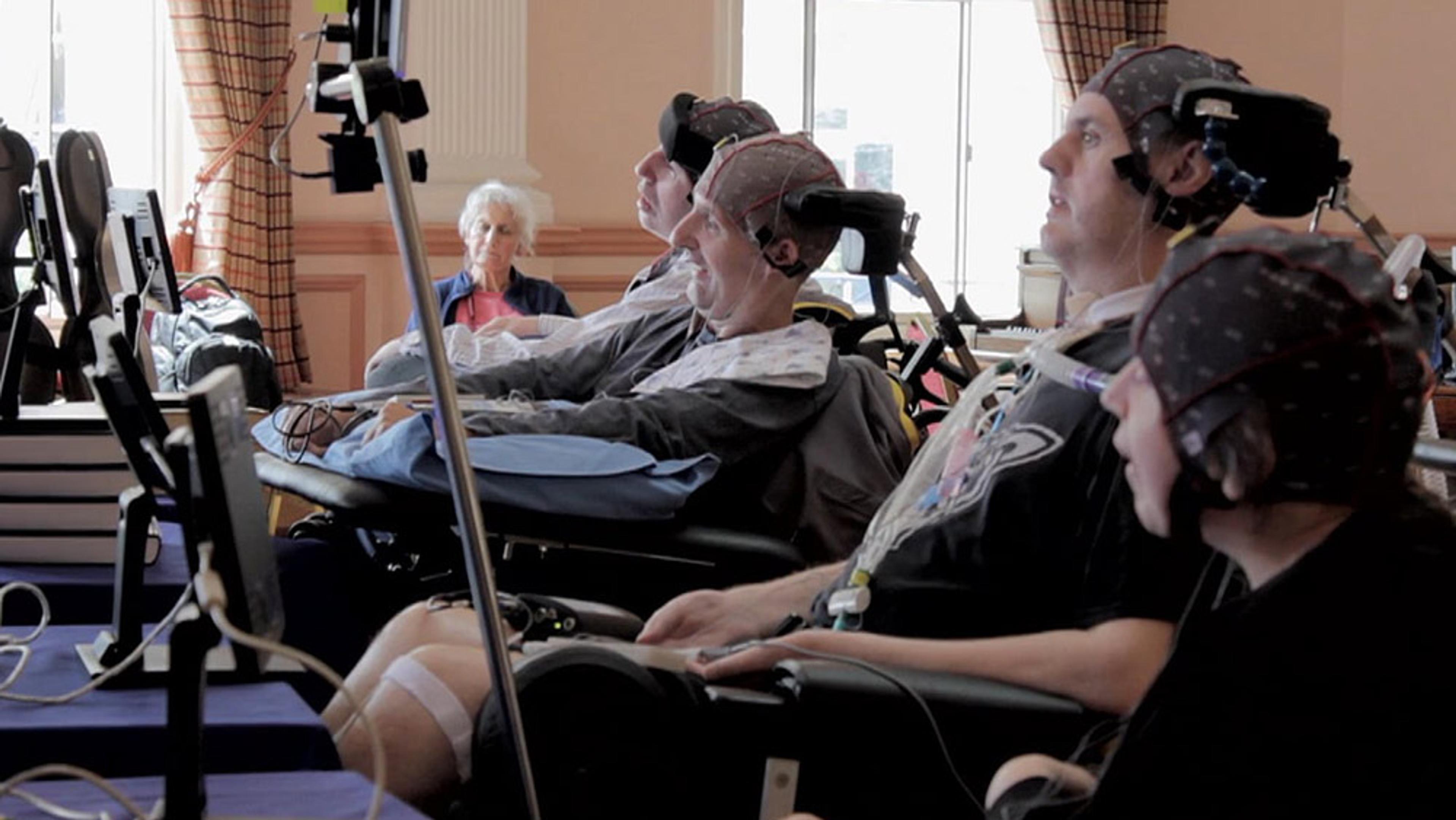 Several disabled people in wheelchairs wearing headgear, using computers in a room.