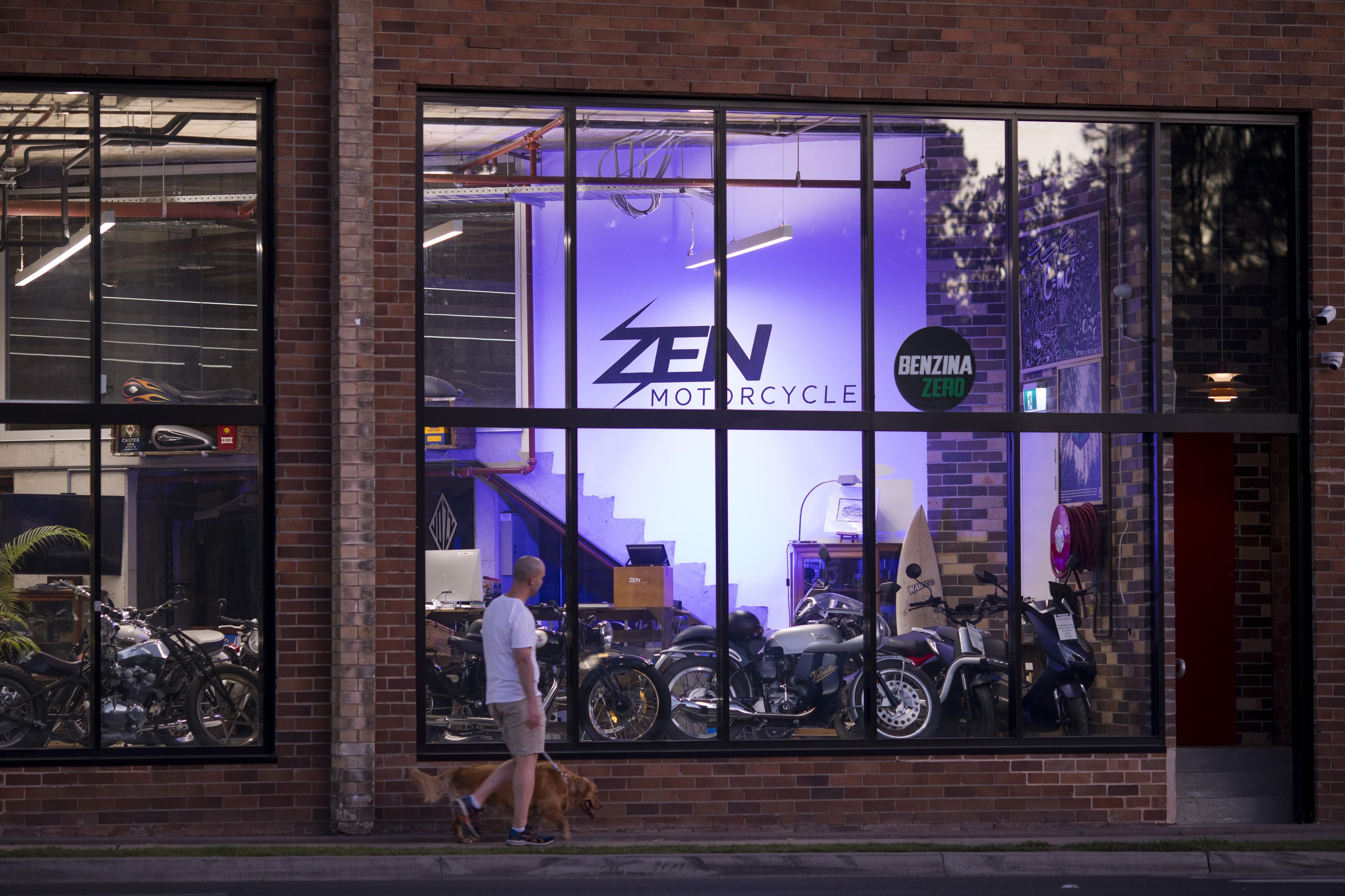 Photo of a man walking a dog past a motorcycle shop with large windows brick exterior and illuminated interior.