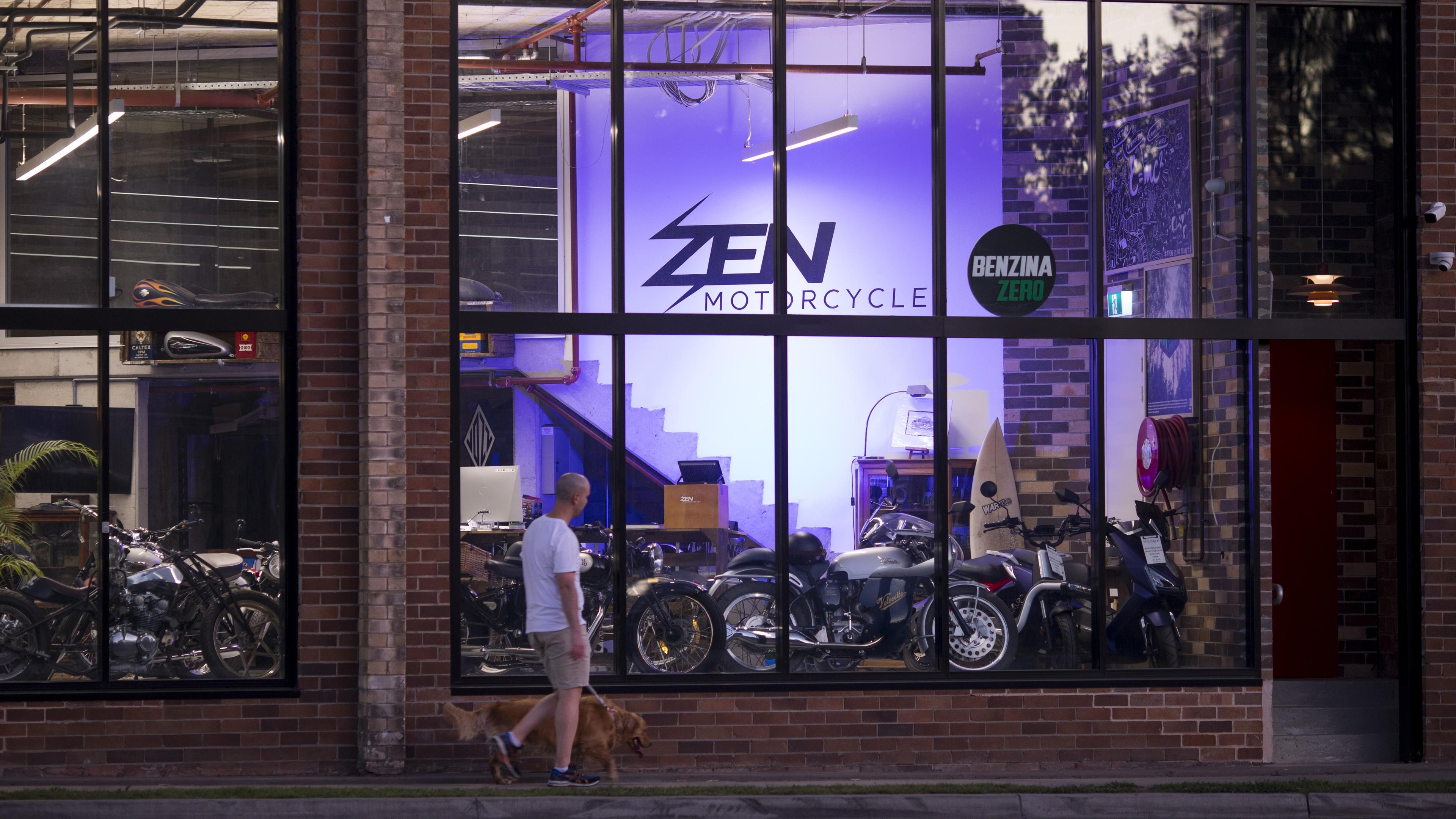 Photo of a man walking a dog past a motorcycle shop with large windows brick exterior and illuminated interior.