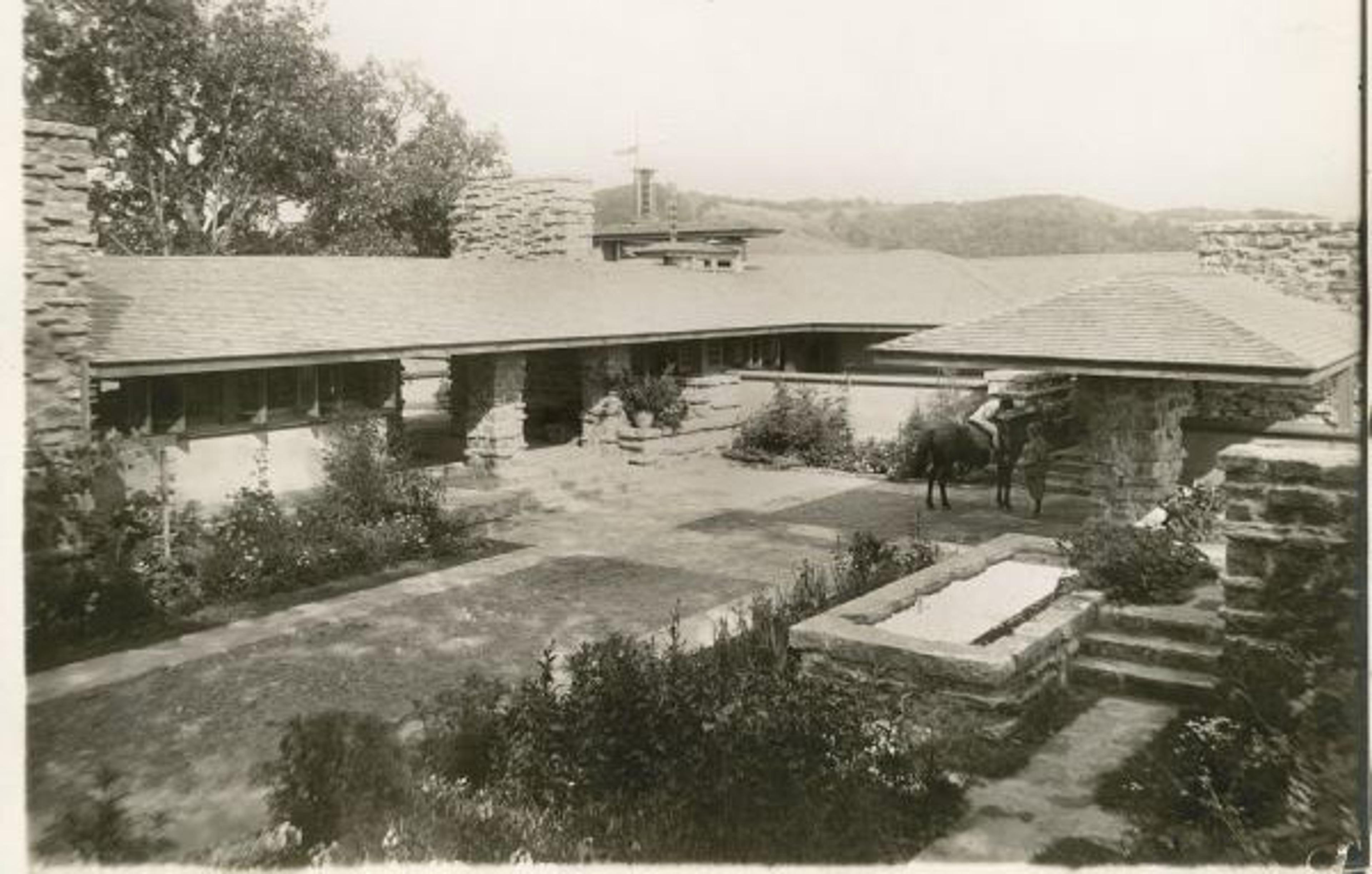 Black and white photo of a large stone house with a horse in the courtyard, surrounded by trees and hills in the background.