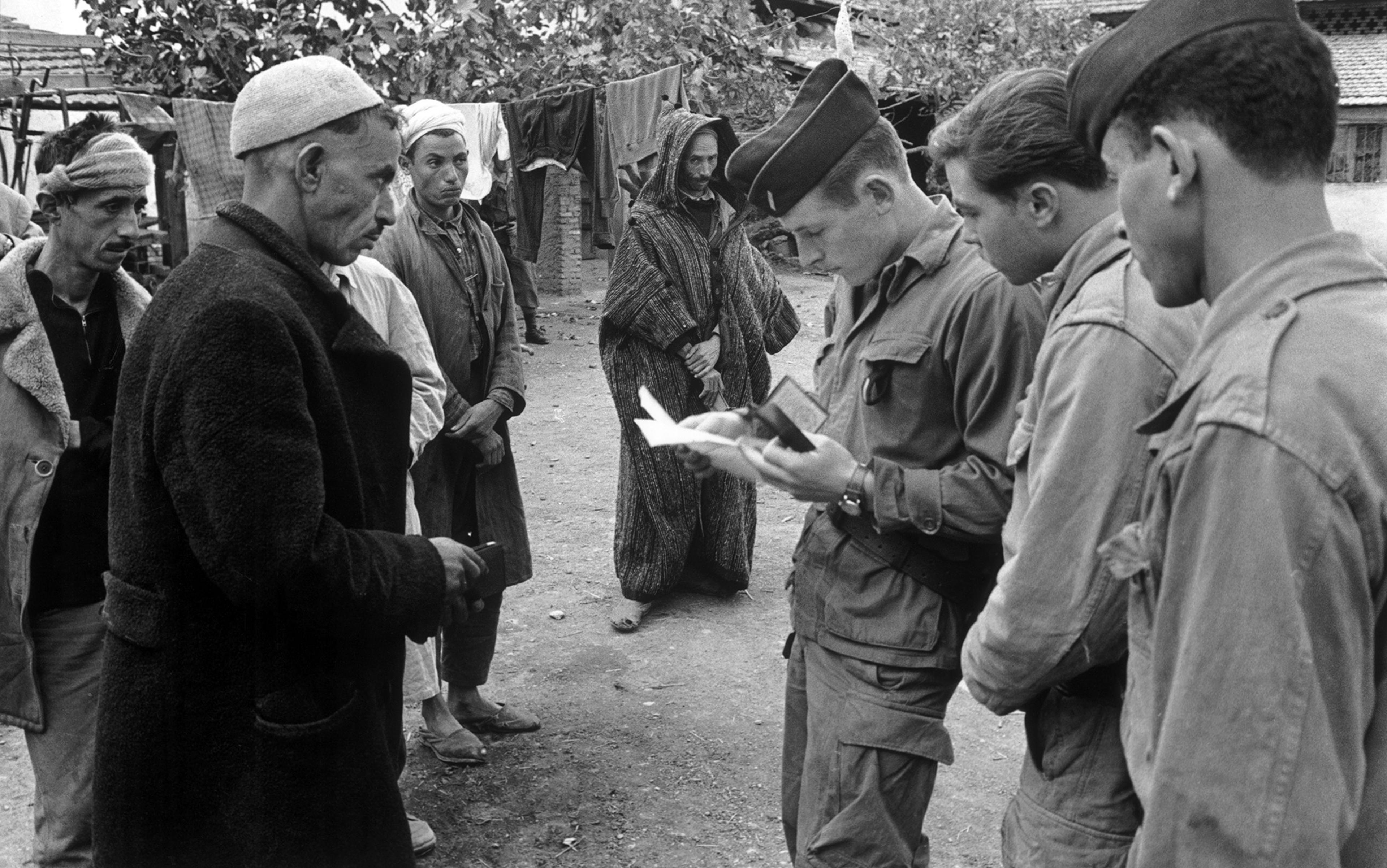 A black-and-white photo of soldiers in uniform checking documents of several men standing outdoors, with laundry hanging in the background.