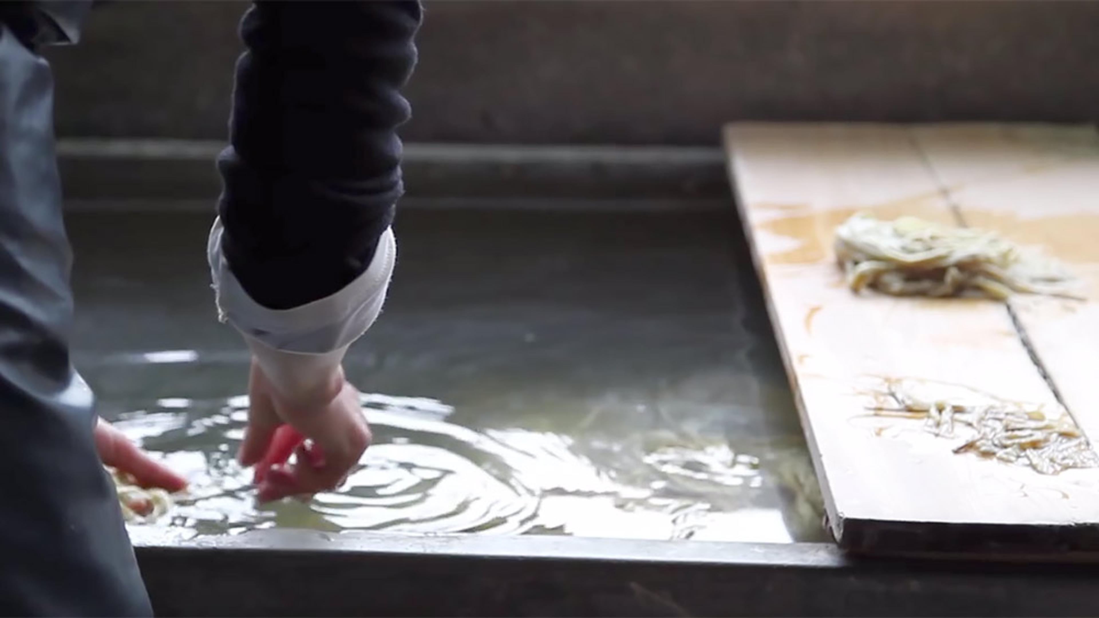 A person with their hand in a water tank next to a wooden board with wet material. The scene suggests a paper-making process.