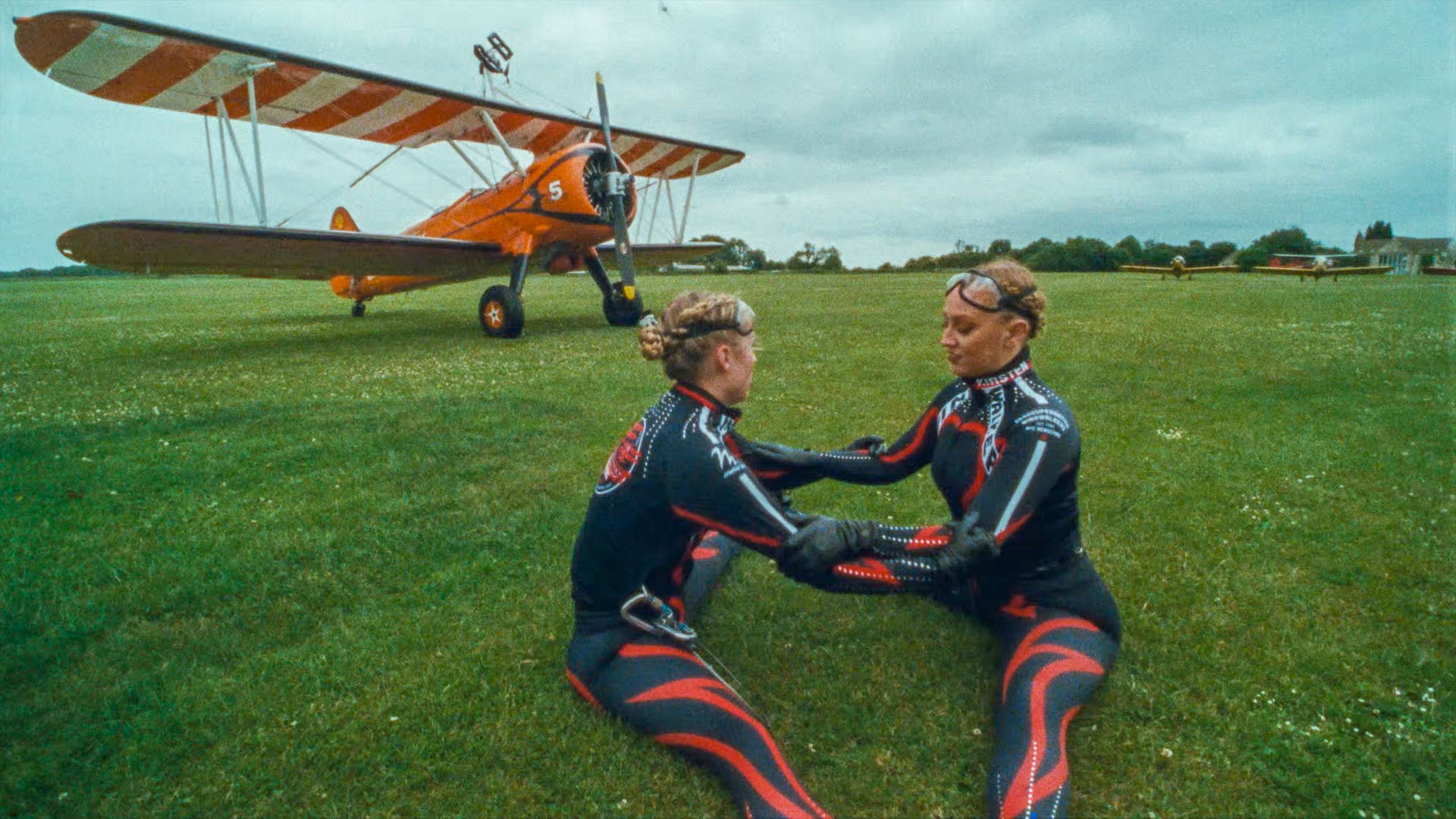 Photo of two women in black and red jumpsuits sitting on grass in front of an orange biplane on a cloudy day.