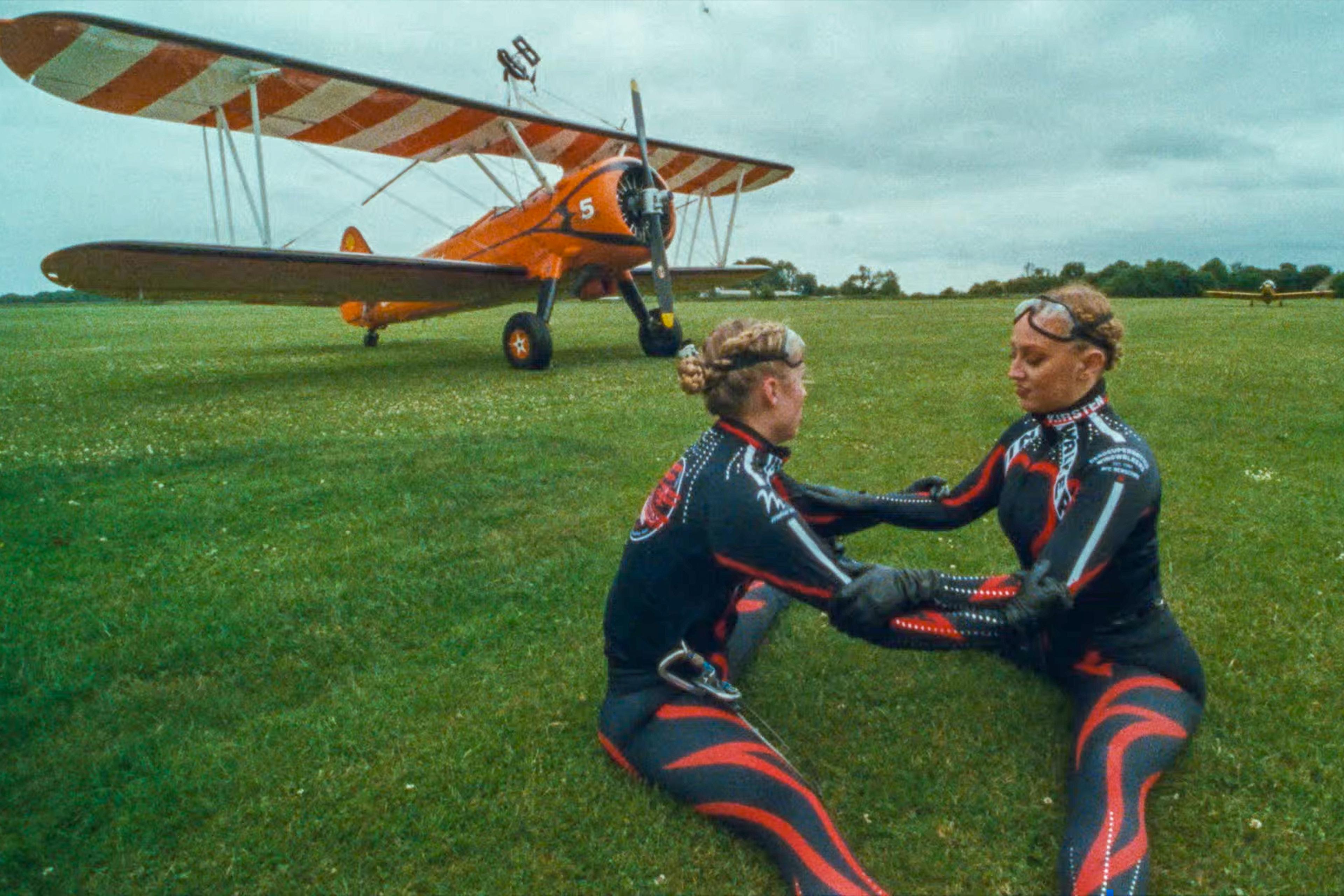 Photo of two women in black and red jumpsuits sitting on grass in front of an orange biplane on a cloudy day.