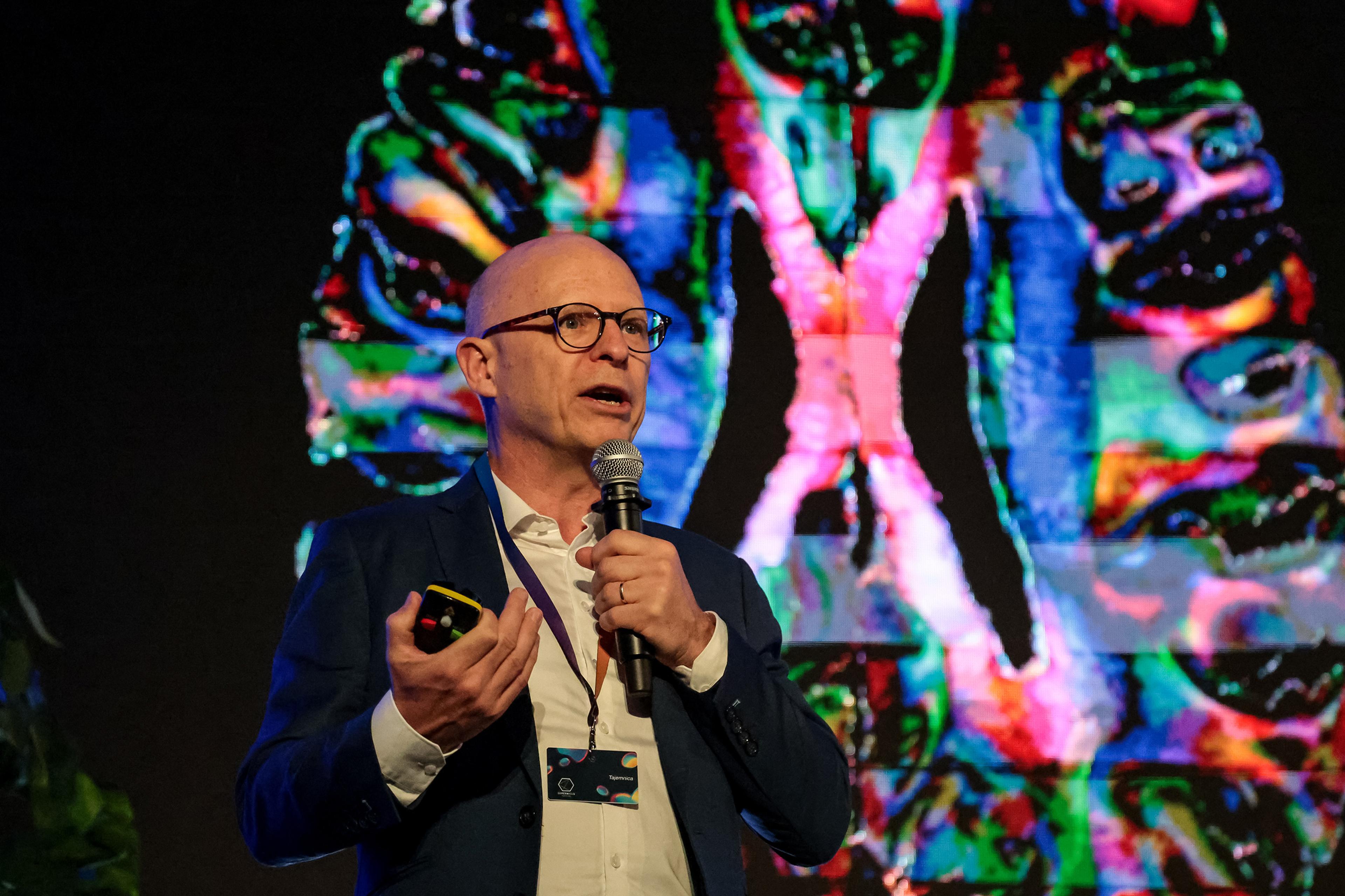 A man in a suit speaks into a microphone in front of a vibrant, abstract digital background; photo.