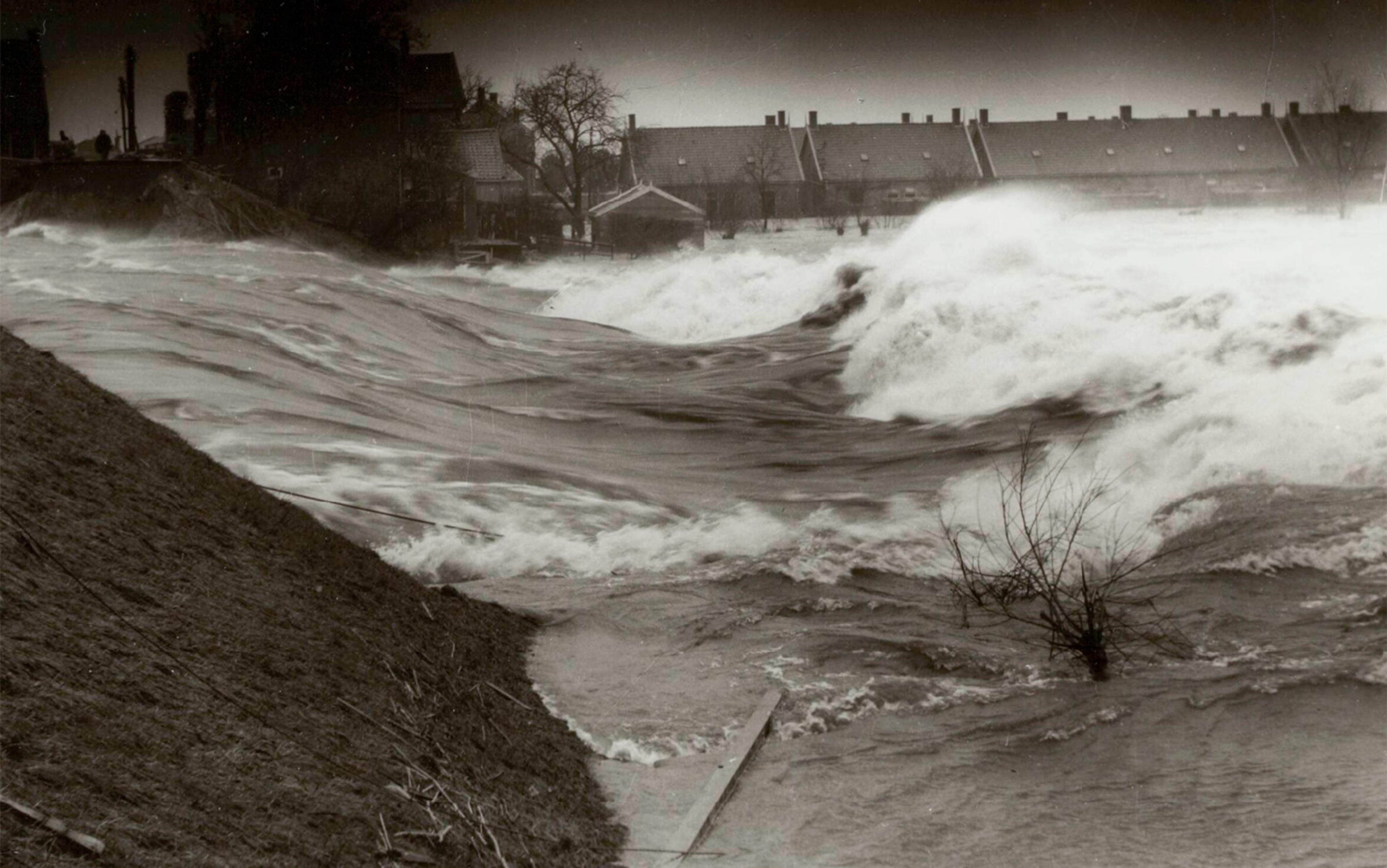 Black and white photograph depicts a flood with rising water levels in a residential area. Strong currents and waves are visible, and houses in the background are partially submerged. Floodwater covers much of the landscape, with a lone tree and partial wooden structure in the foreground.