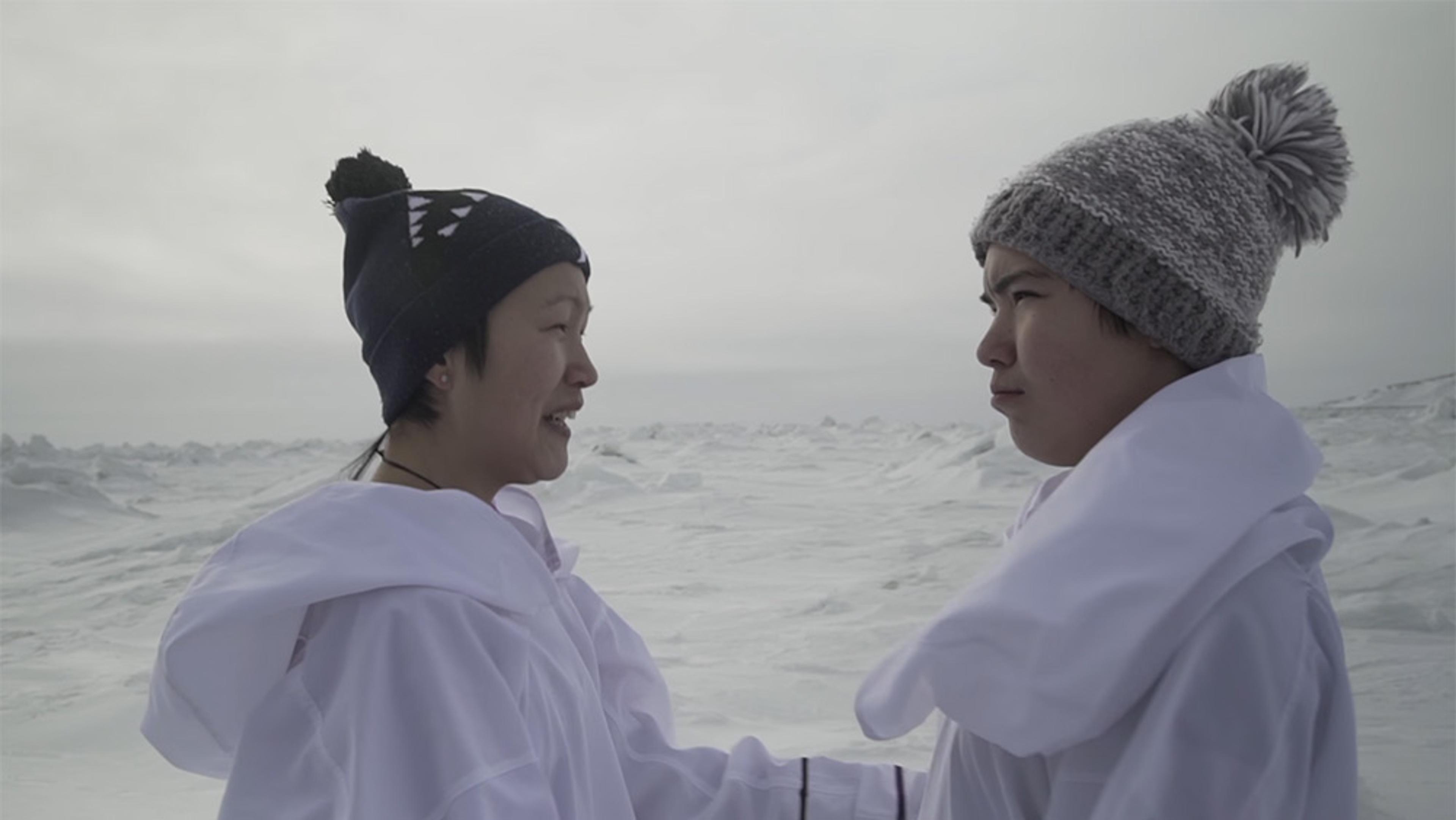 Two people in white robes and winter hats standing in a snowy landscape, facing each other, one smiling, the other serious.